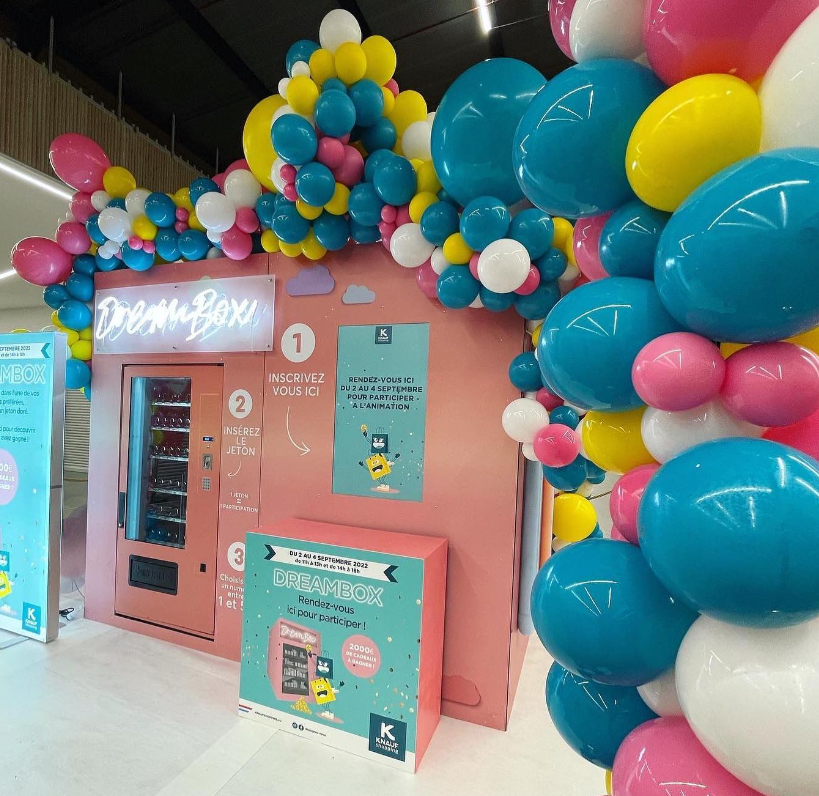 Decorative display with pink background, colorful balloons in blue, pink, yellow, and white, and a vending machine labeled 'Dream Box' for registration and participation in an event.