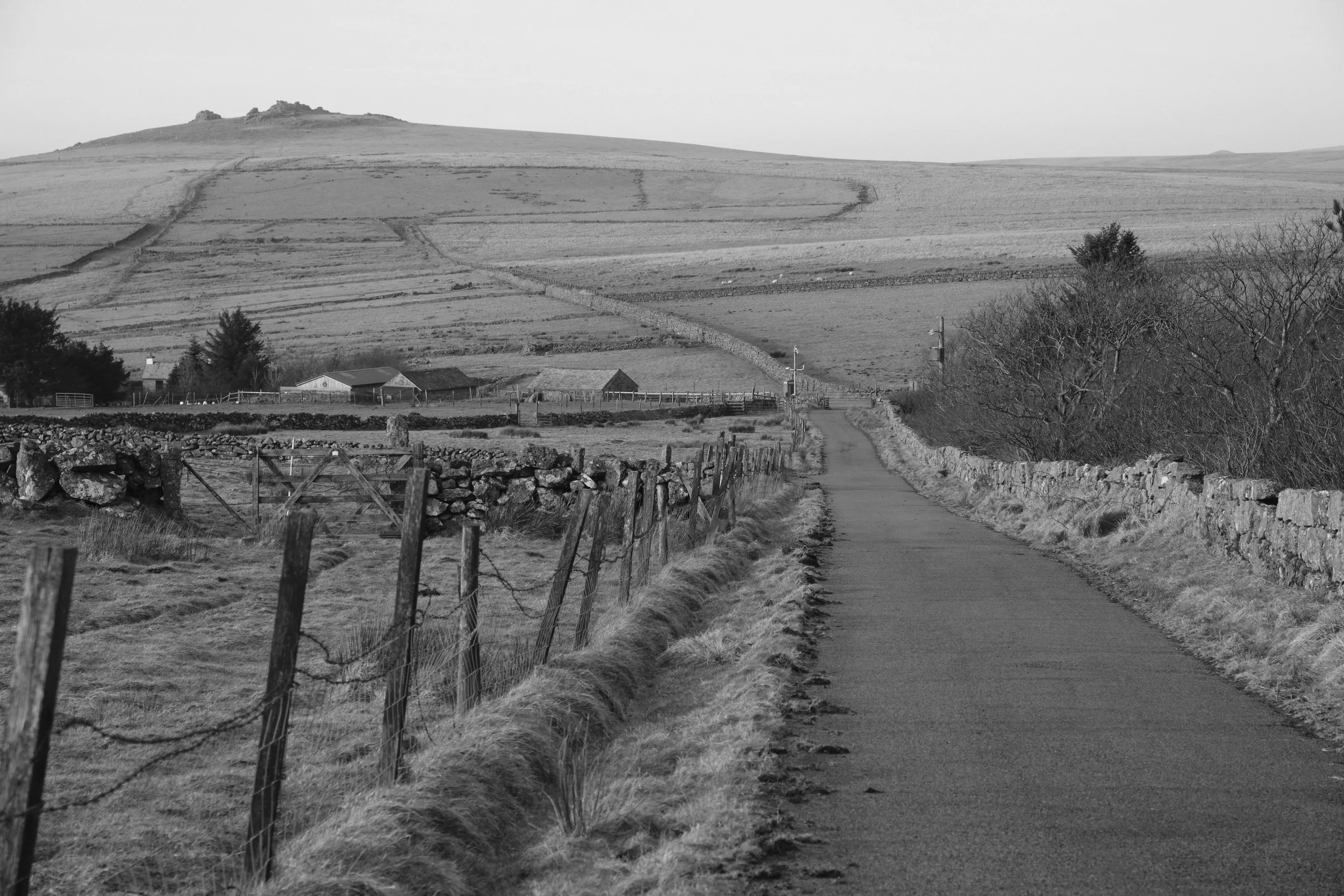 A black and white photo of a rural landscape with a narrow road, dry stone walls on each side, and fields on hills in the background.