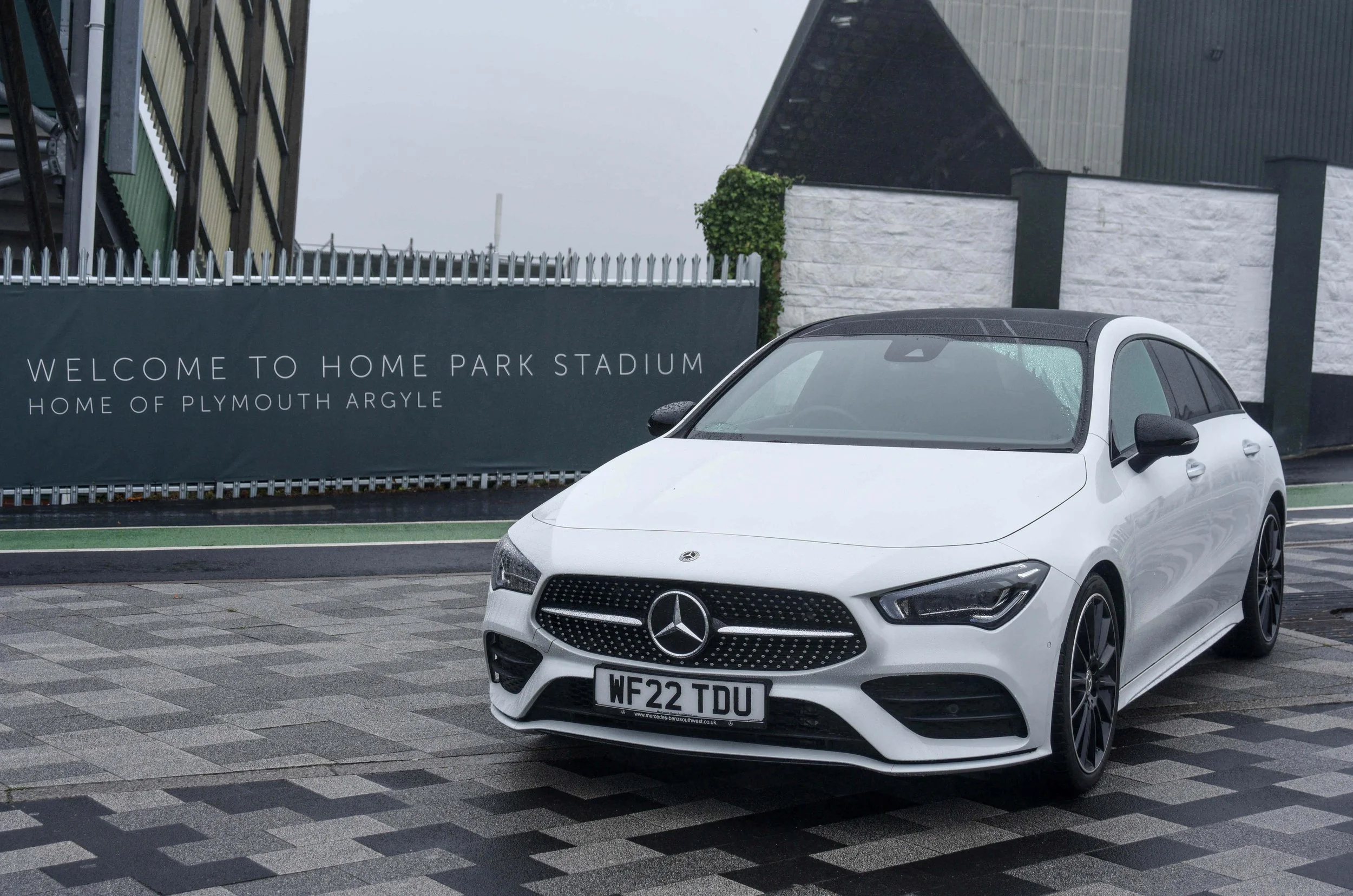 A white Mercedes-Benz is parked on a wet, patterned pavement in front of a sign reading "Welcome to Home Park Stadium, Home of Plymouth Argyle," under a gray, overcast sky