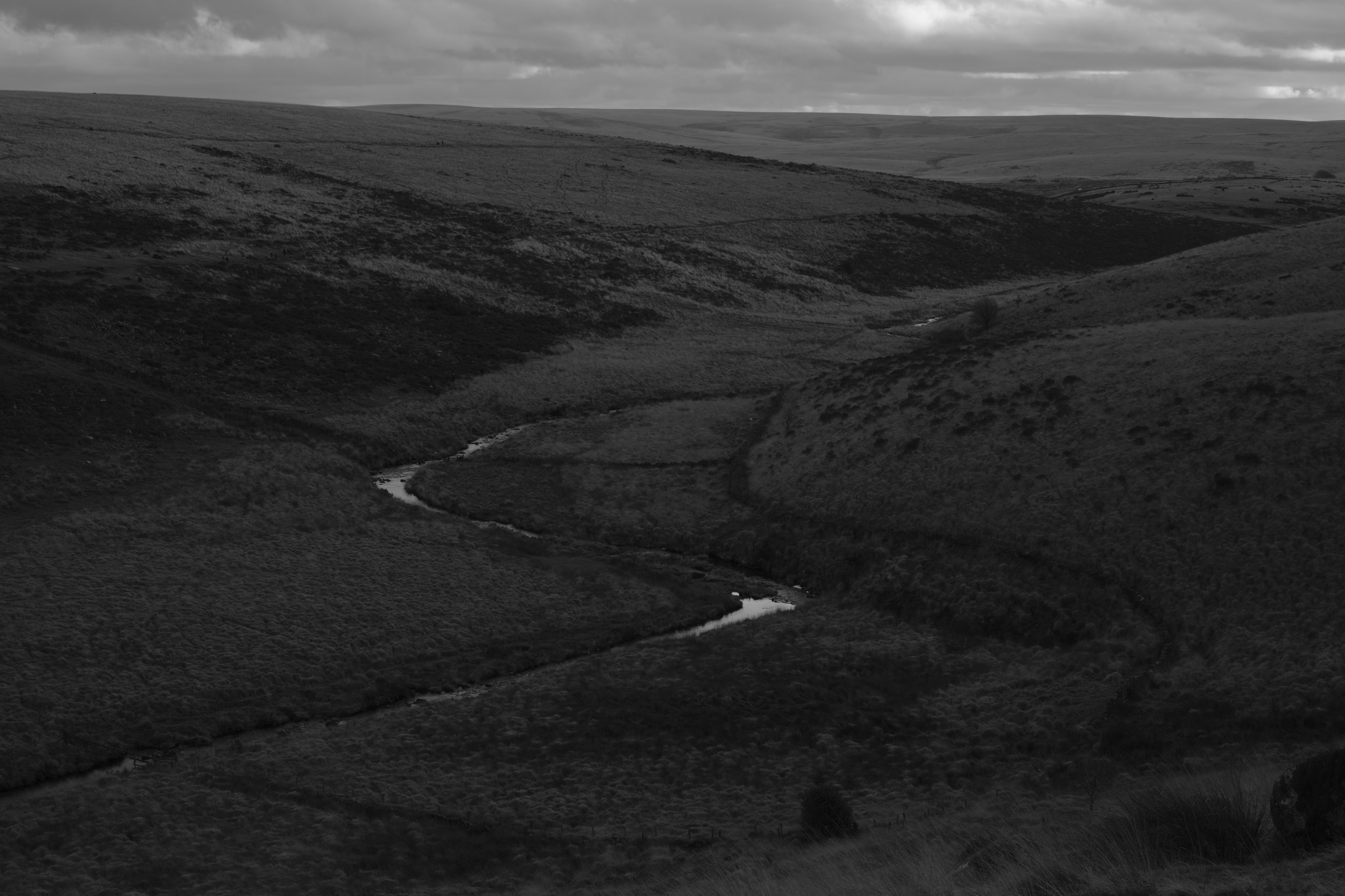 Black and white landscape showing a winding river through rolling hills under an overcast sky. The scene is tranquil and expansive.