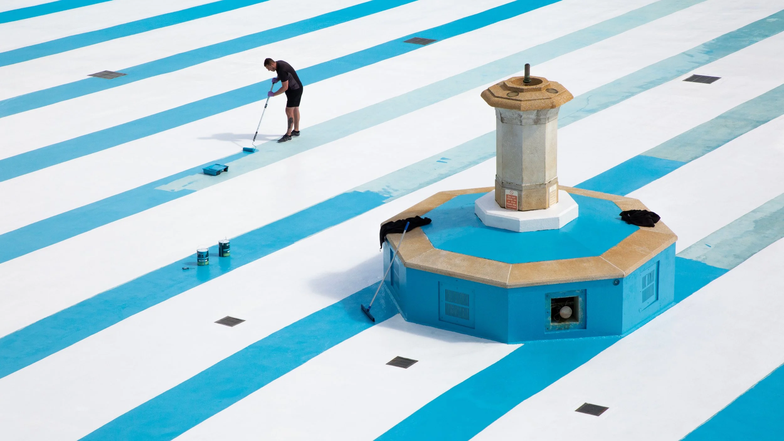 A person paints light blue stripes on an empty, drained swimming pool around a central octagonal fountain. The scene conveys a sense of solitude and focus.