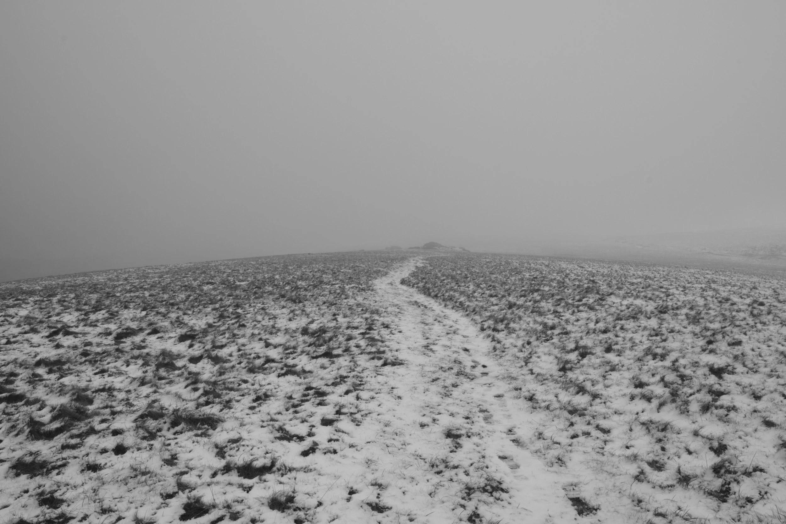 Black and white photograph of Yes Tor from Matt Marshall's Dystopia. The landscape is covered in snow with patches of grass sticking out and footprints visible along a main path down the middle of the image. Set in misty conditions