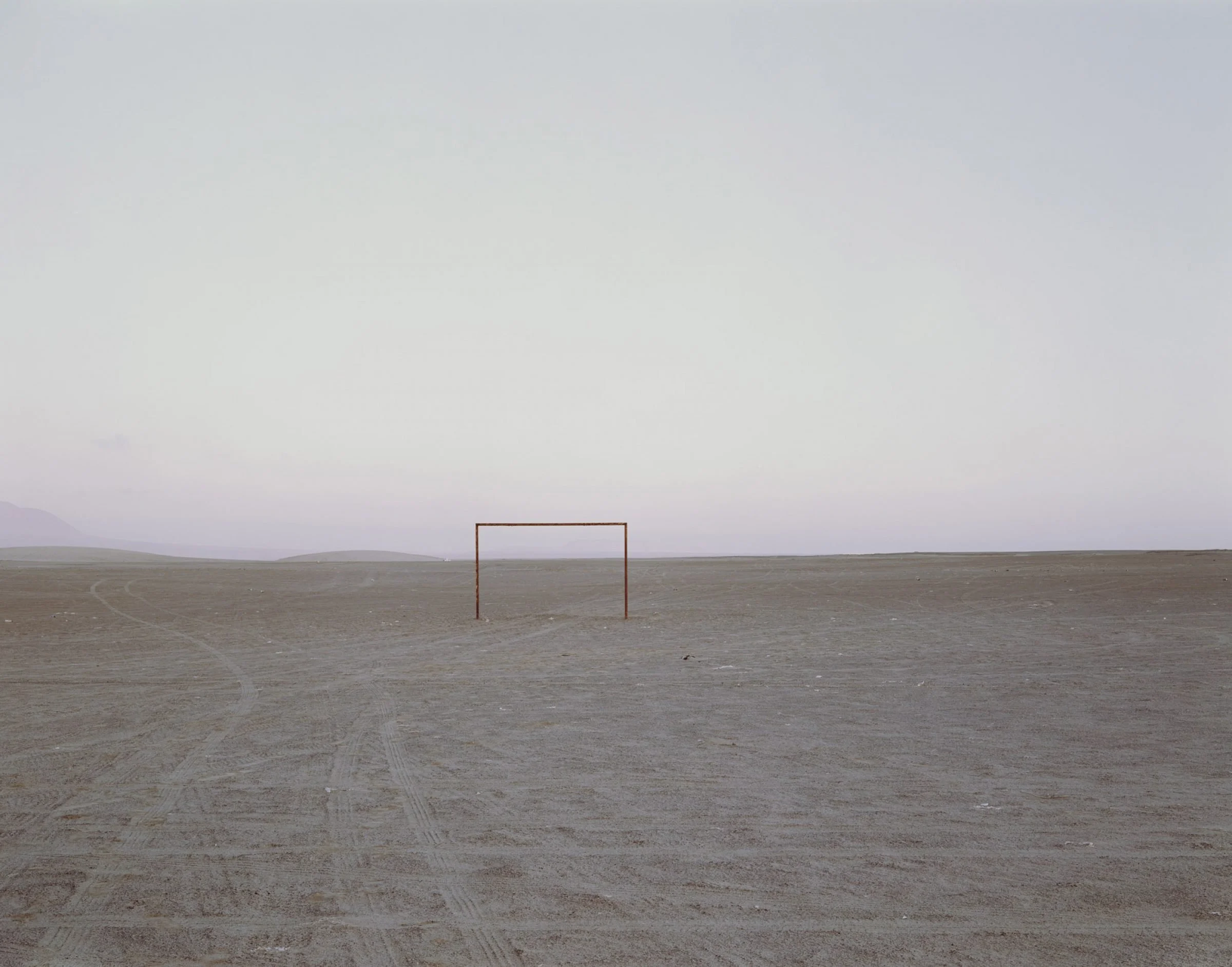 Colour photograph from Nadav Kander's God's Country depicting a football goalpost in the middle of a barren land where tyre tracks can be seen to the left of shot.
