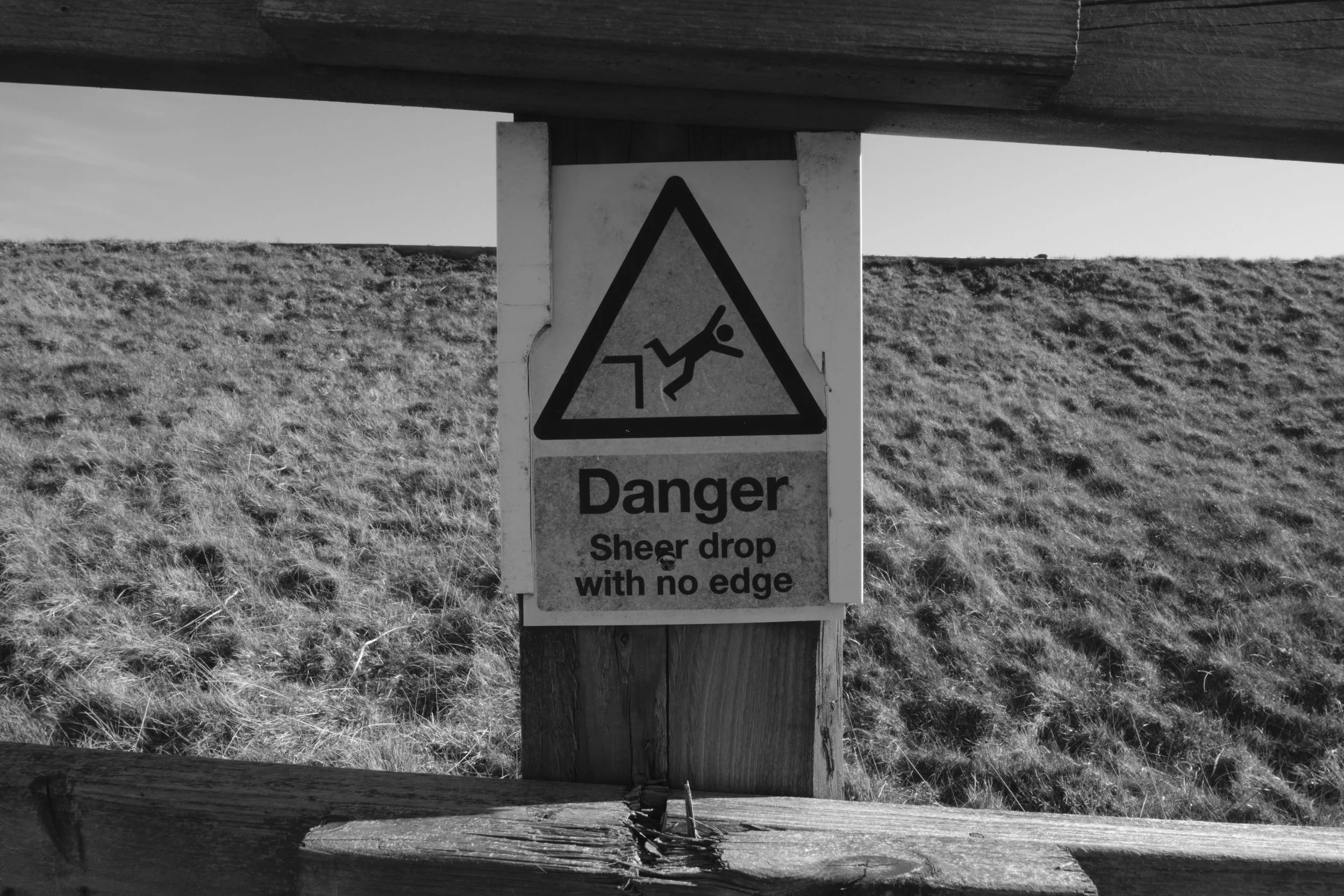 Warning sign showing a person falling from a shear drop with no edge, on a wooden post in an outdoor grassy area.