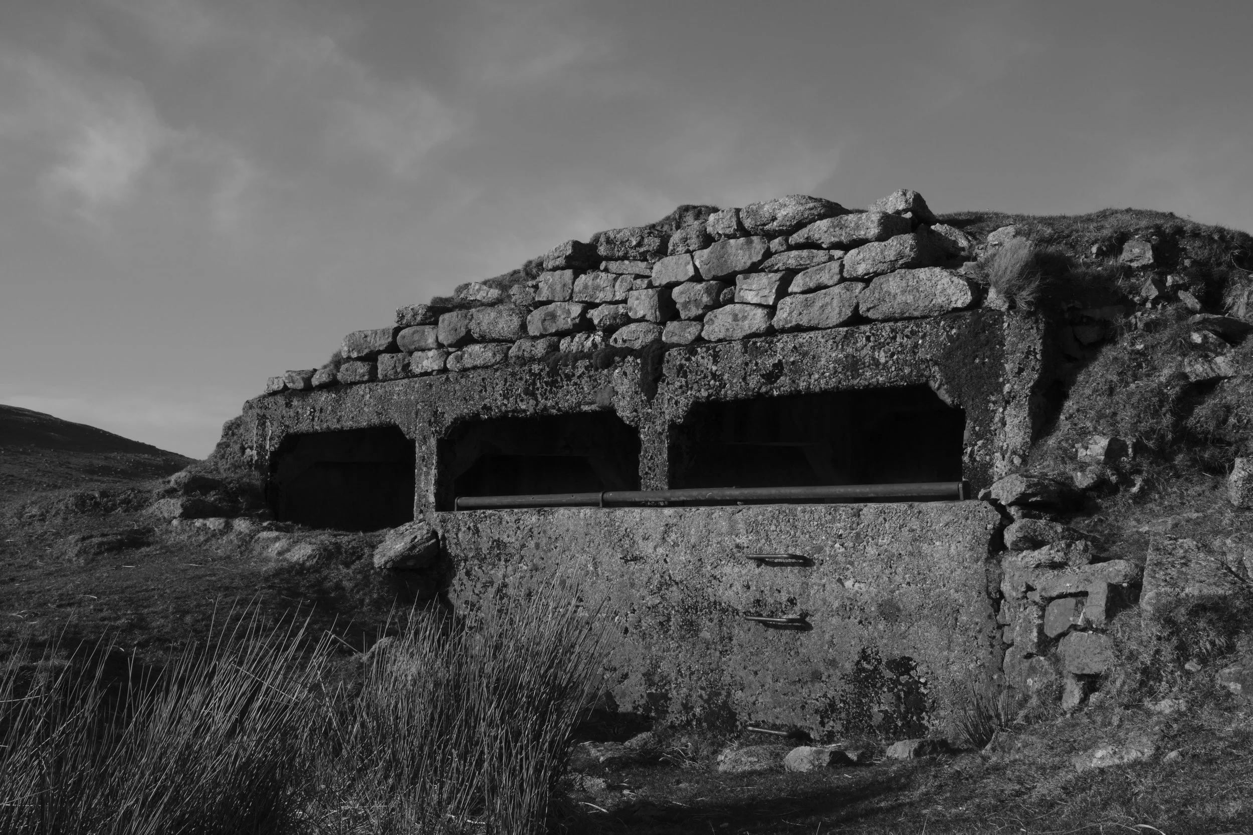 Black and white photograph of an old stone military bunker or defensive structure built into a grassy hillside, featuring rough stacked stone walls, three dark rectangular openings and set in a remote rural landscape under a cloudy sky.