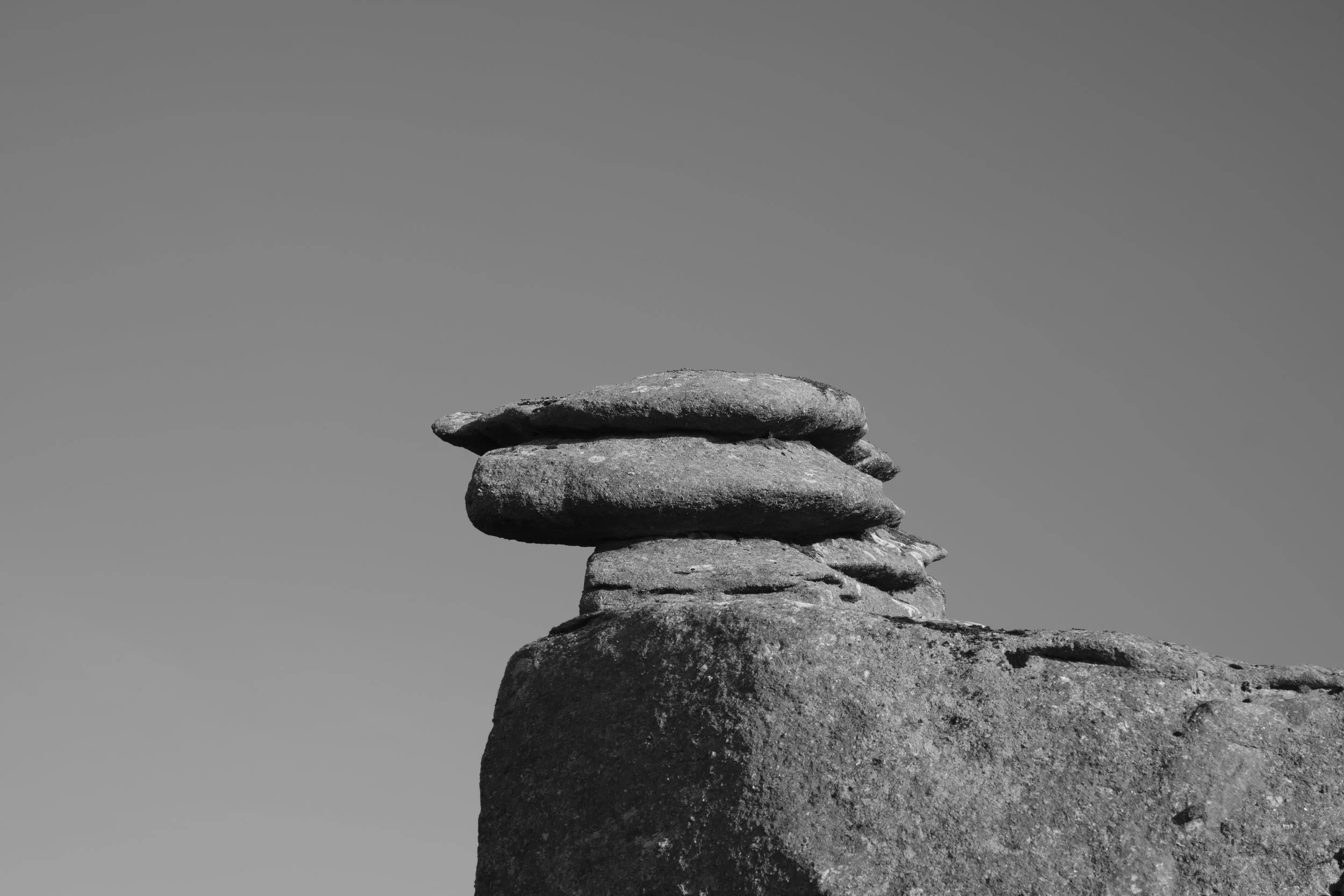Black and white image of a stack of flat stones balanced on a rock against a clear sky. The scene conveys simplicity and calmness.