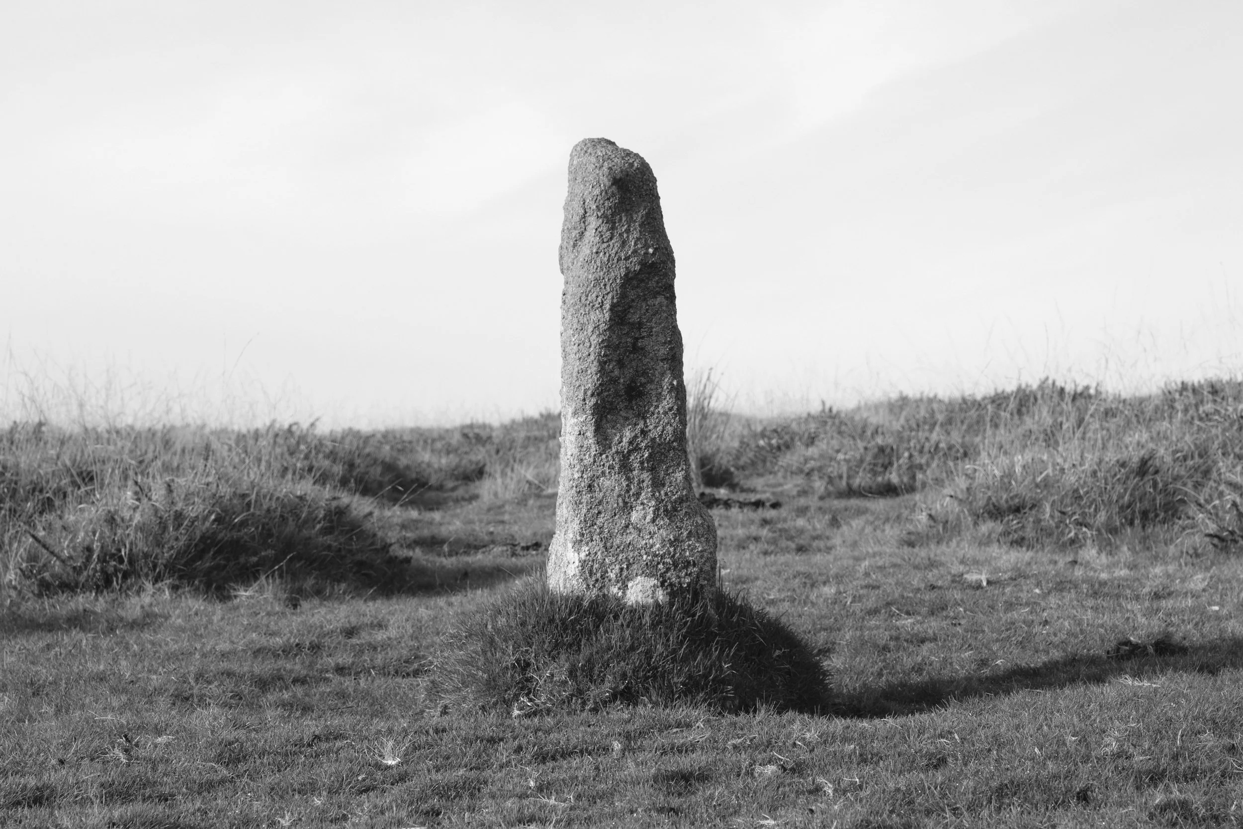 A solitary standing stone in a grassy landscape with gentle slopes, under a cloudy sky, in black and white.