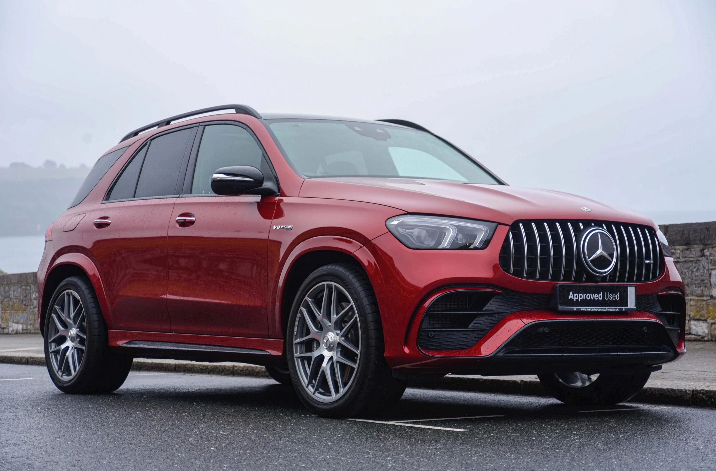 A red Mercedes-Benz SUV is parked on a wet road beside a stone wall, with water and hills visible in the background.
