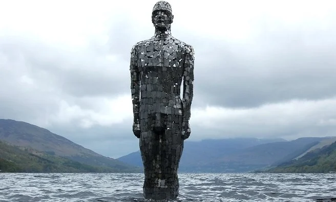 Large iron human figure sculpture standing waist-deep in a wide lake, facing forward with arms at sides, constructed from stacked rectangular metal plates. The dark metallic statue contrasts with rippling water in the foreground and misty mountains.