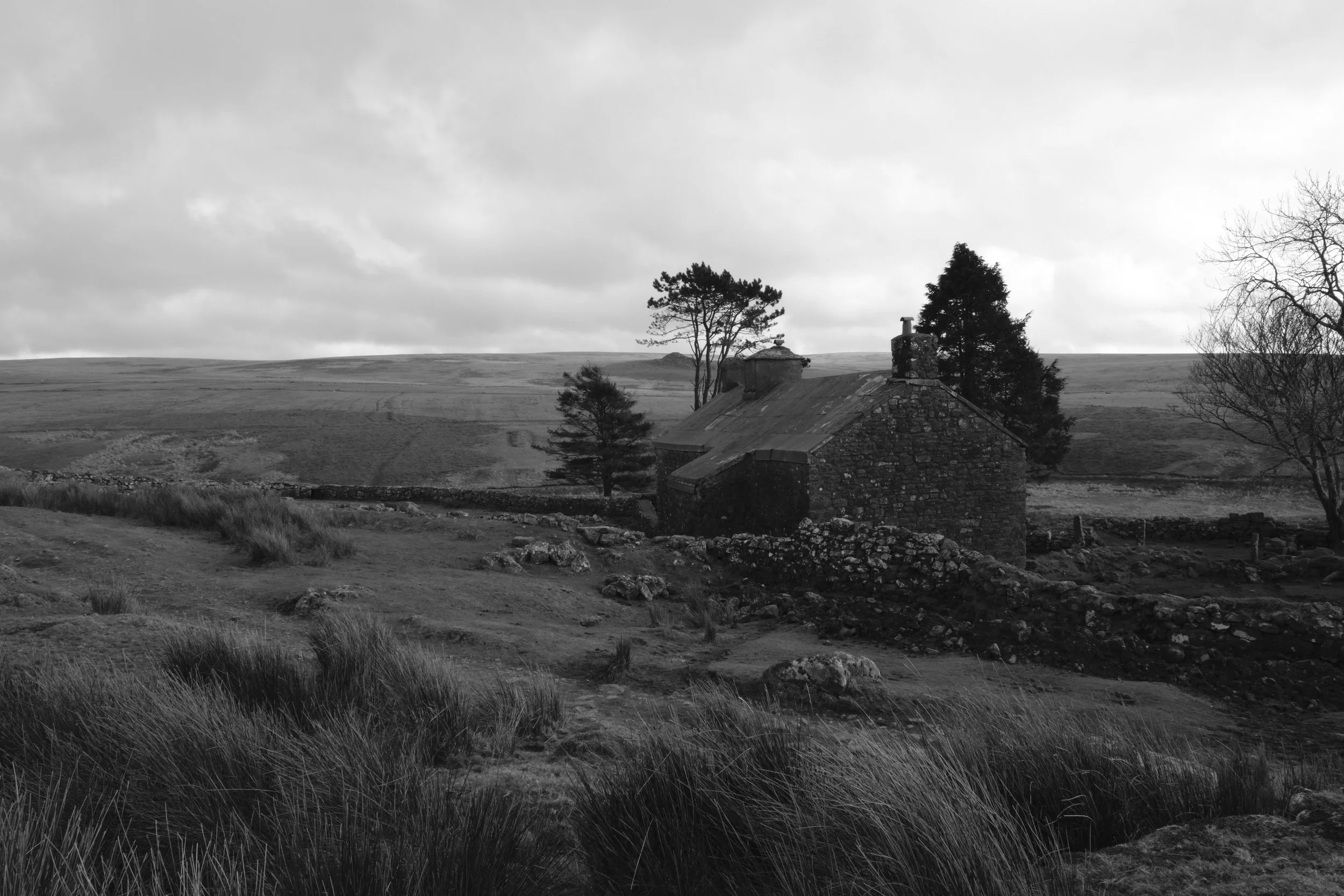 An old stone cottage in a rural landscape with rolling hills, trees, and a cloudy sky, in black and white.