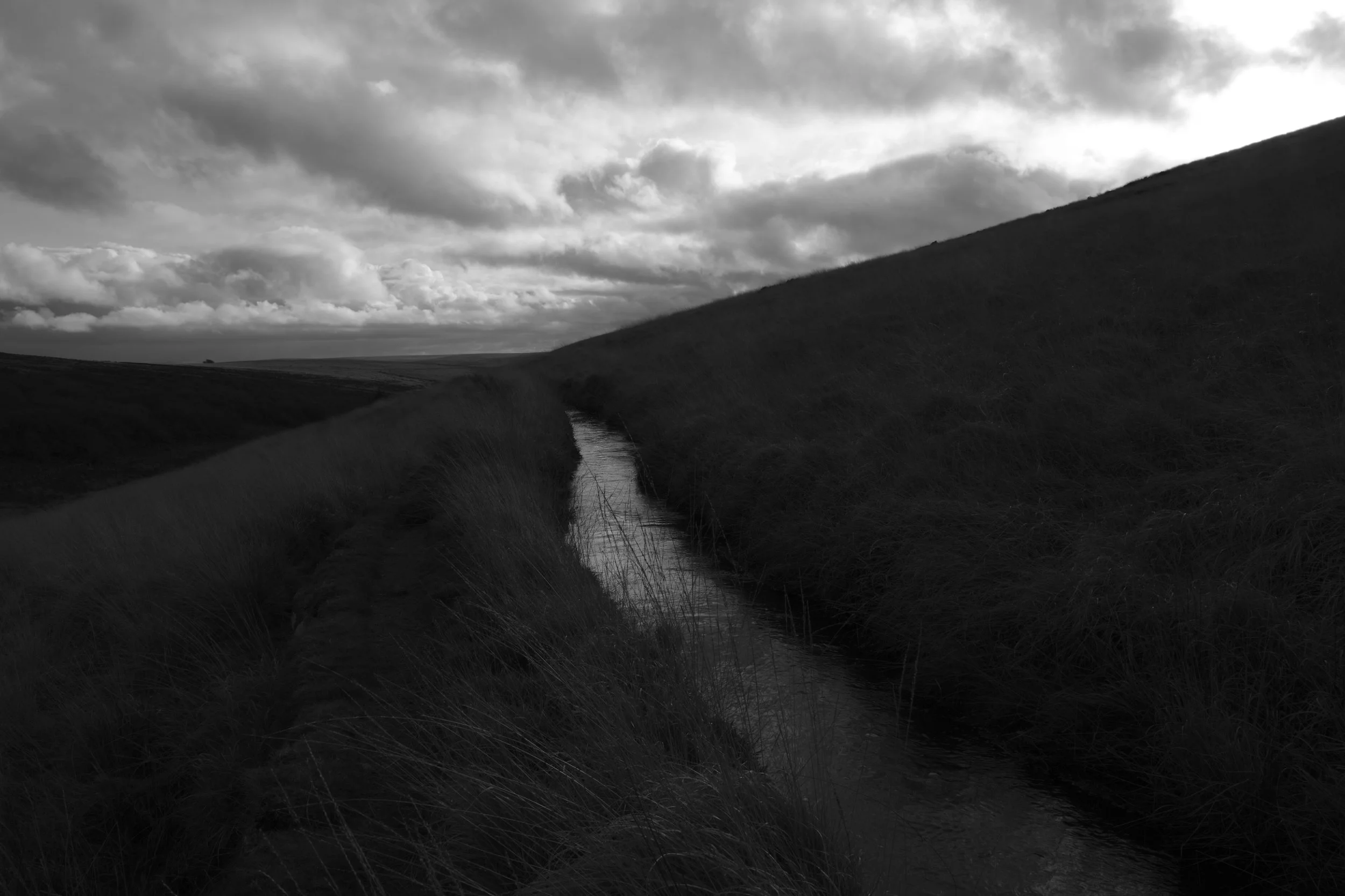 A dark, moody landscape featuring a narrow stream flowing between grassy hills under a cloudy sky. The scene conveys a sense of solitude and tranquility.