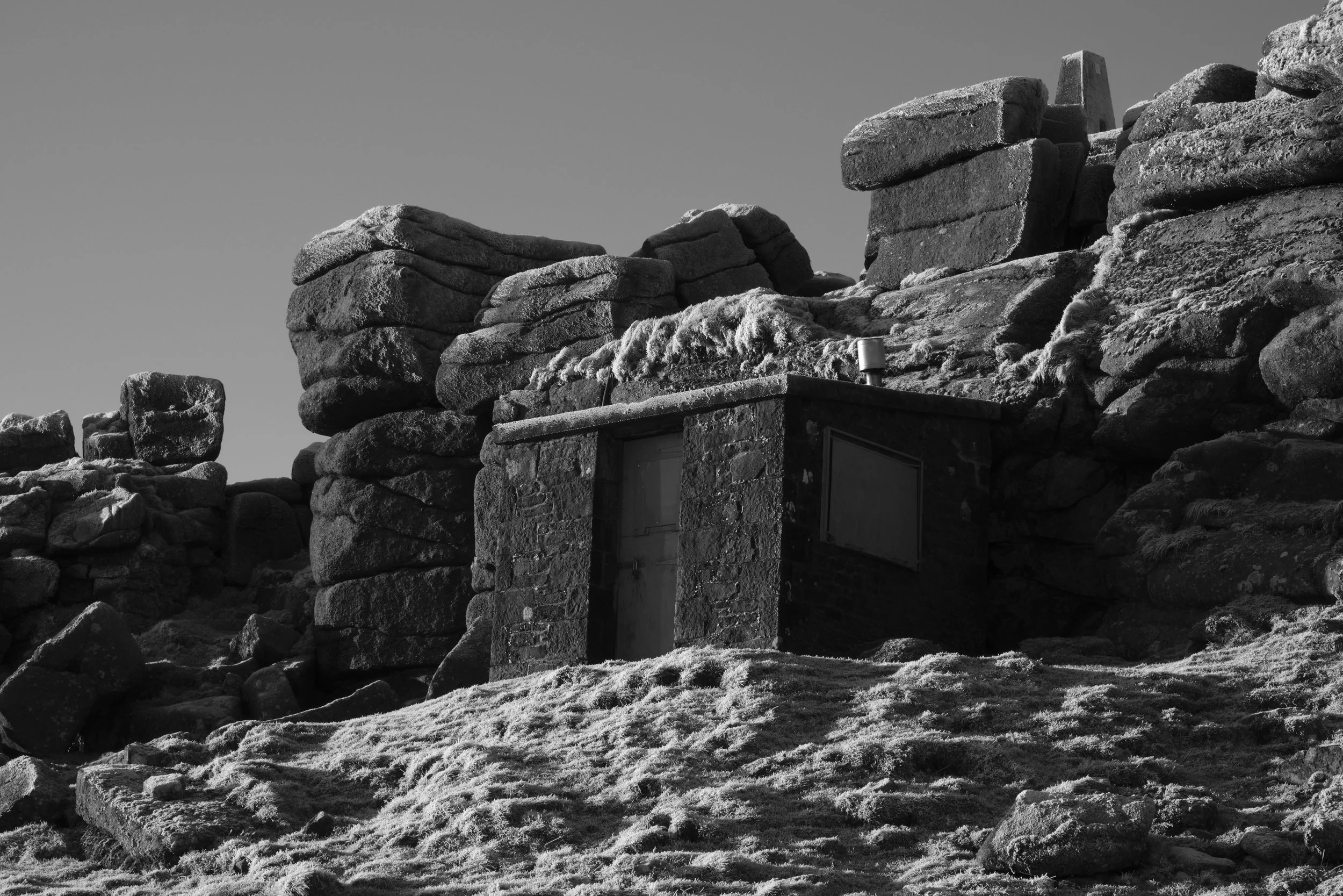 Black and white image of a small stone building nestled among large, rugged rocks with a clear sky above. The scene feels isolated and serene.