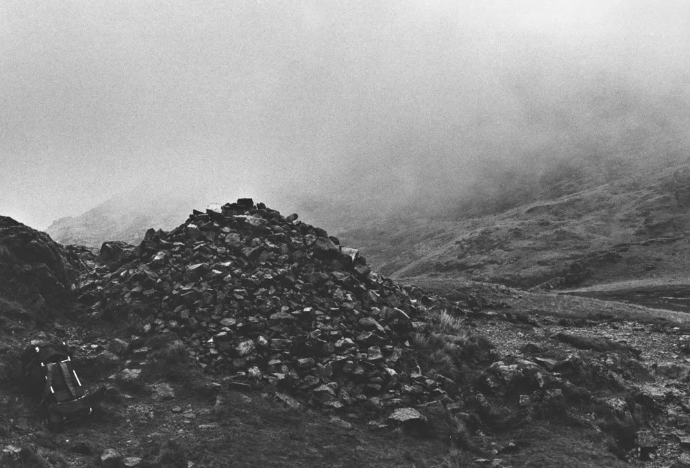 Black and white photograph from Richard Long of stones piles on top of each other with a backpack to the left of the image and mountain terrain obscured by foggy conditions.