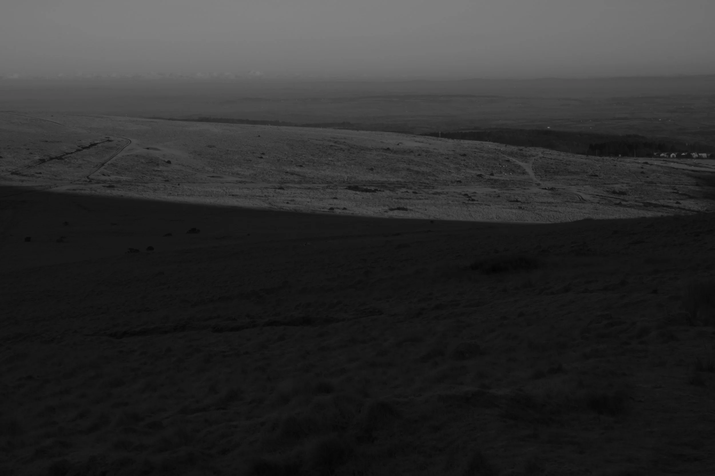 A shadowy, black-and-white image of a barren, expansive landscape. The foreground is dark, contrasting with the sunlit background, creating a stark, moody atmosphere.