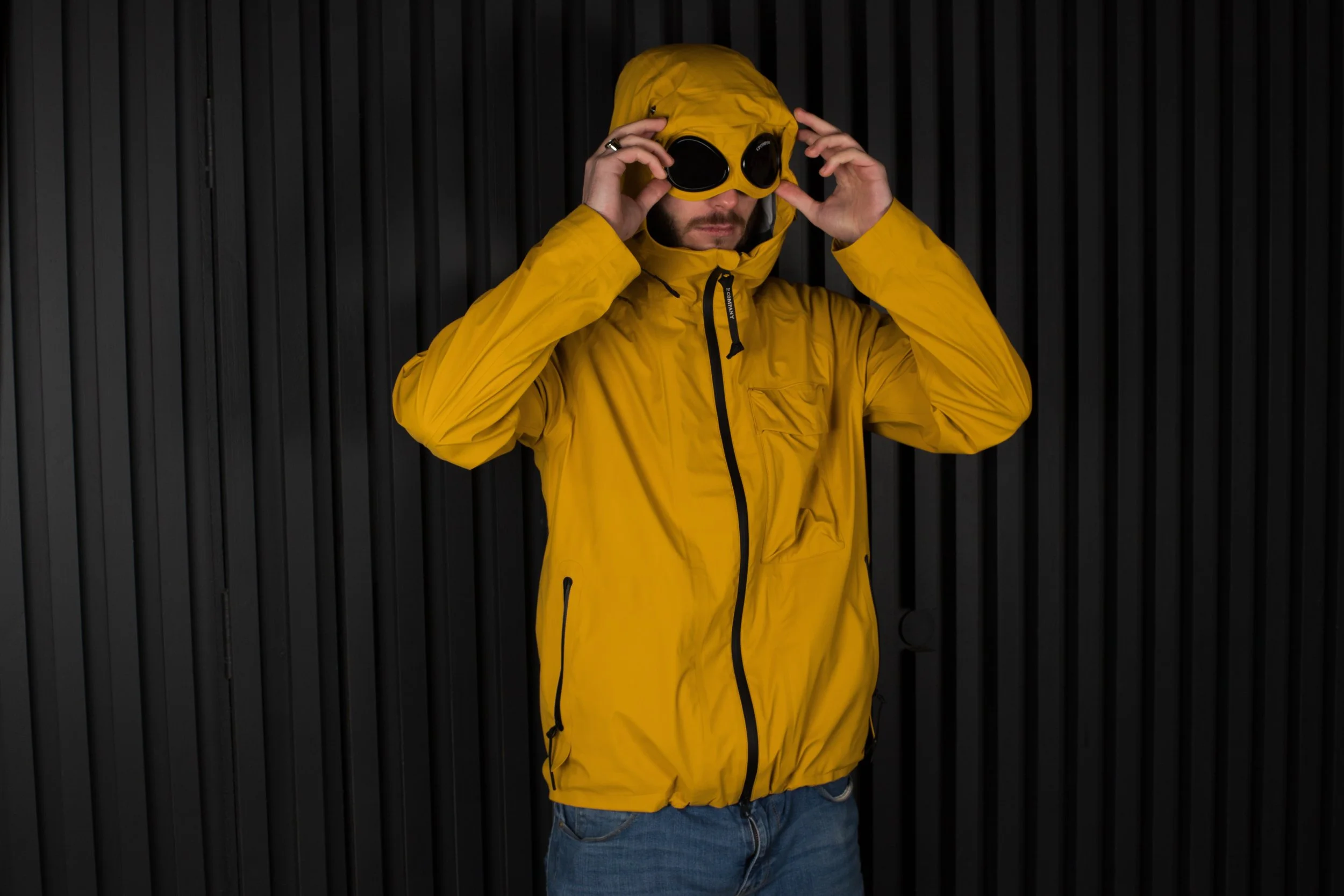 Person in a bright yellow jacket adjusts round goggles on a hoodie. The background is a dark, ribbed surface, creating a bold contrast.