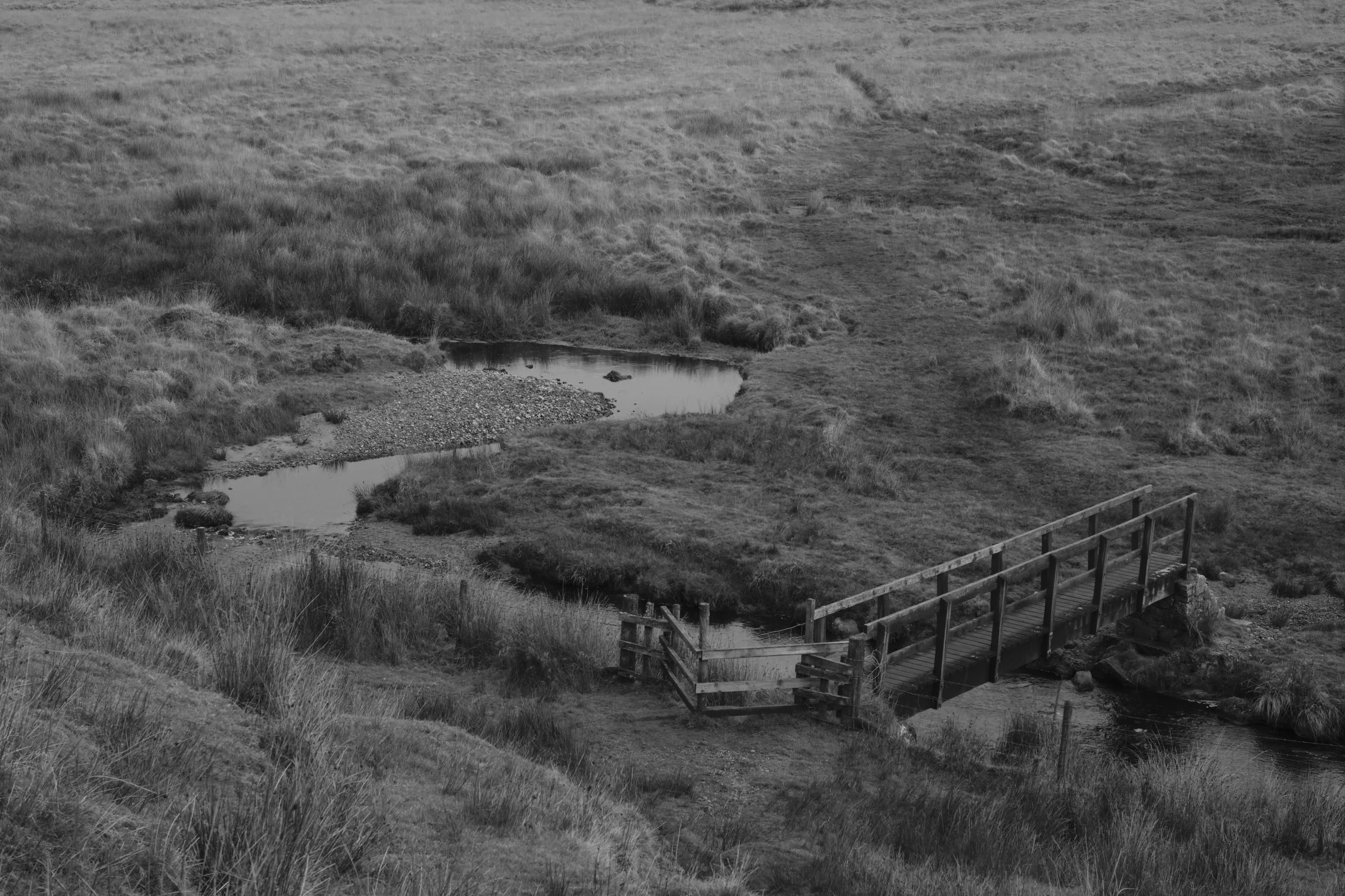 A black and white photo of a small bridge over a stream in a grassy field with hills in the background.