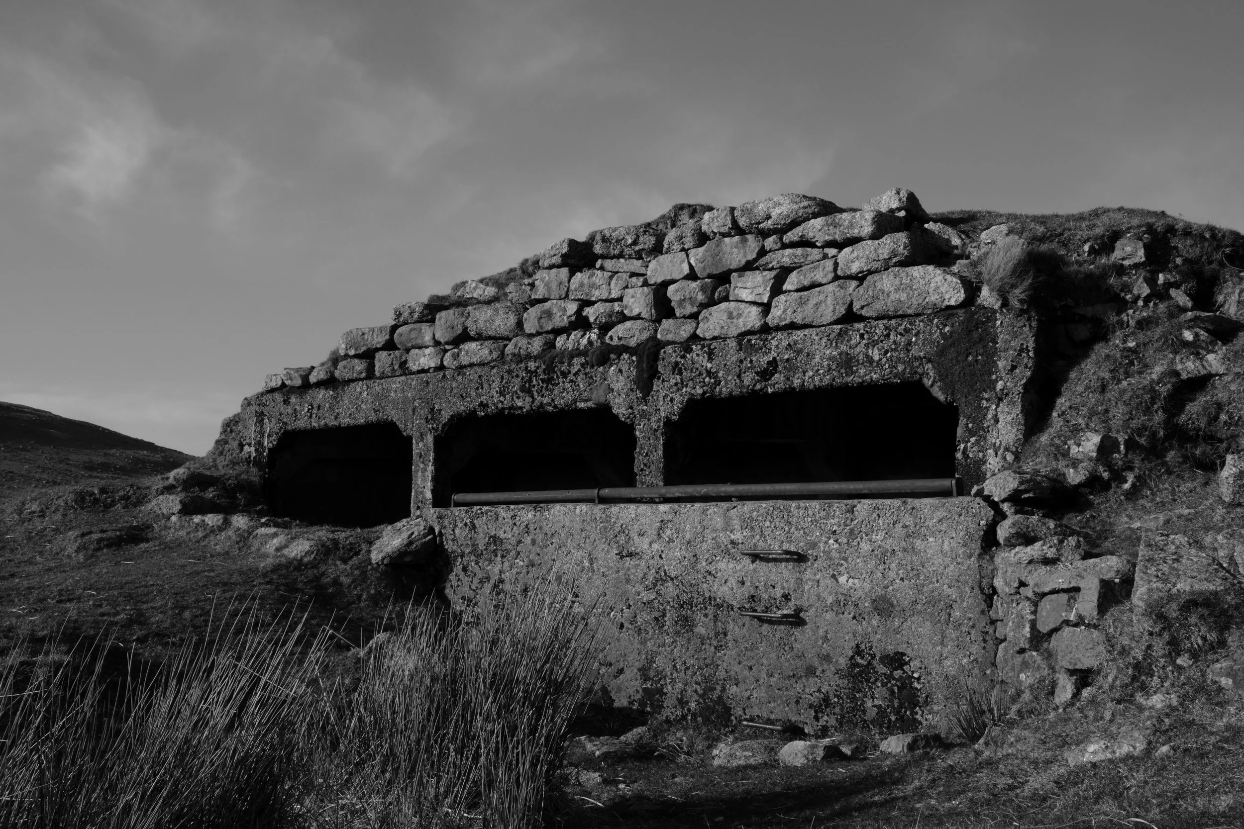 A monochrome image of a weathered stone bunker nestled in a grassy hill. The rough texture and shadowy openings evoke a sense of historical solitude.