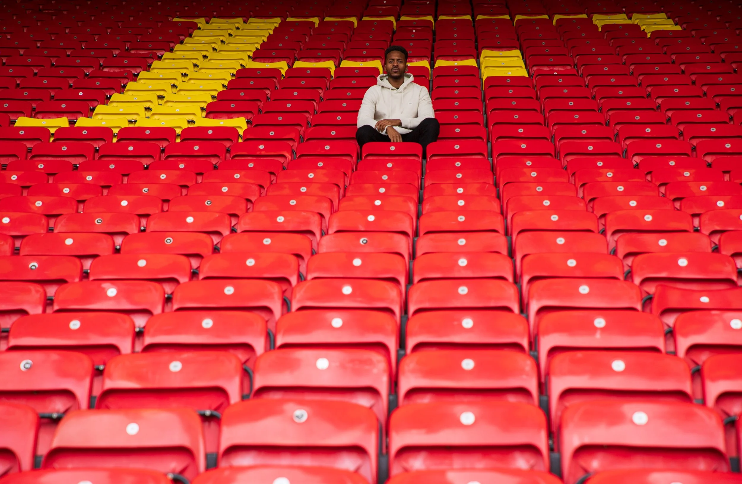 Man seated alone in a stadium on red seats, with yellow seats forming a pattern. The setting is empty and the mood is contemplative.