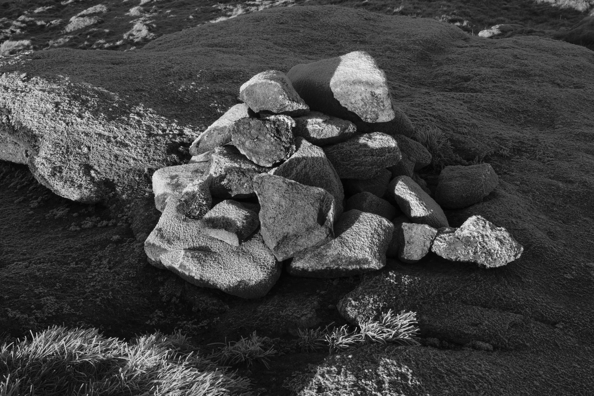 A monochrome image features a small, neatly stacked cairn on a rocky surface. The scene conveys a sense of calm and solitude in a rugged landscape.