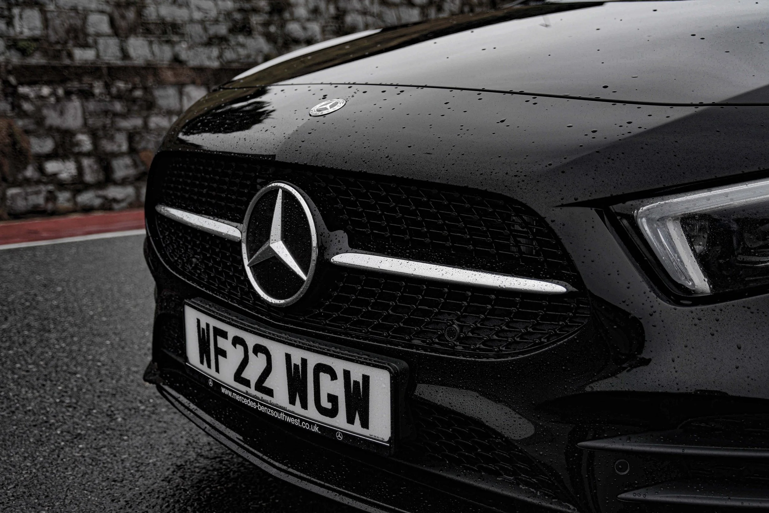 Close-up of a sleek black car's front, featuring a prominent Mercedes emblem on a rain-speckled grille, with a UK license plate below. Mood: elegant.