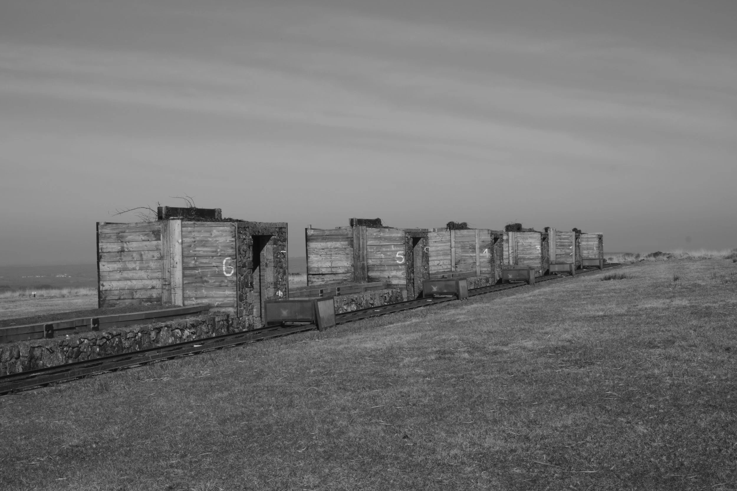 Black and white photo of a row of small wooden huts on a train track, numbered 6, 5, 4, and 3, in an open grassy landscape with a cloudy sky.