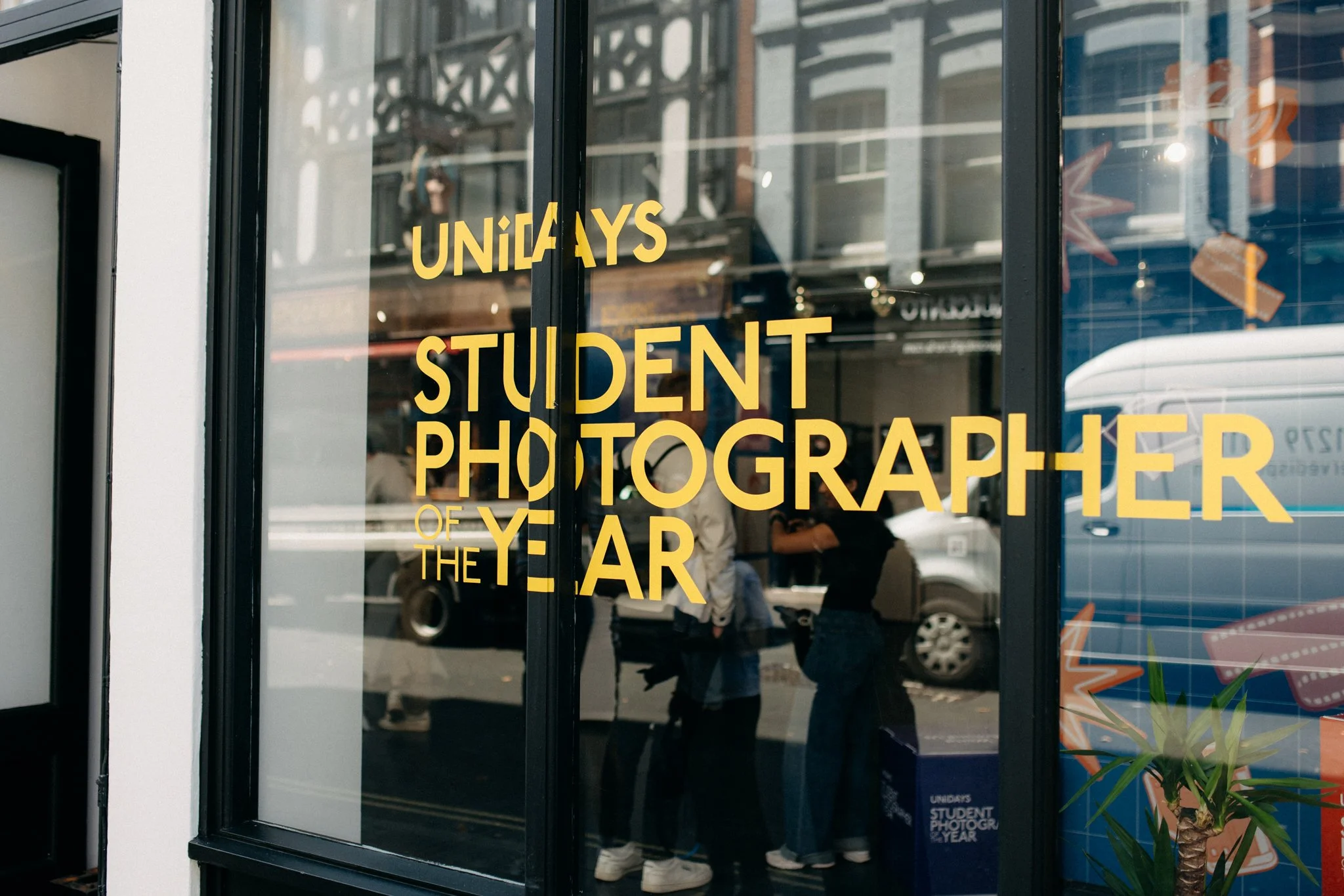 A window with the words "UNiDAYS Student Photographer of the Year" in yellow text; people and street reflections are visible in the glass.