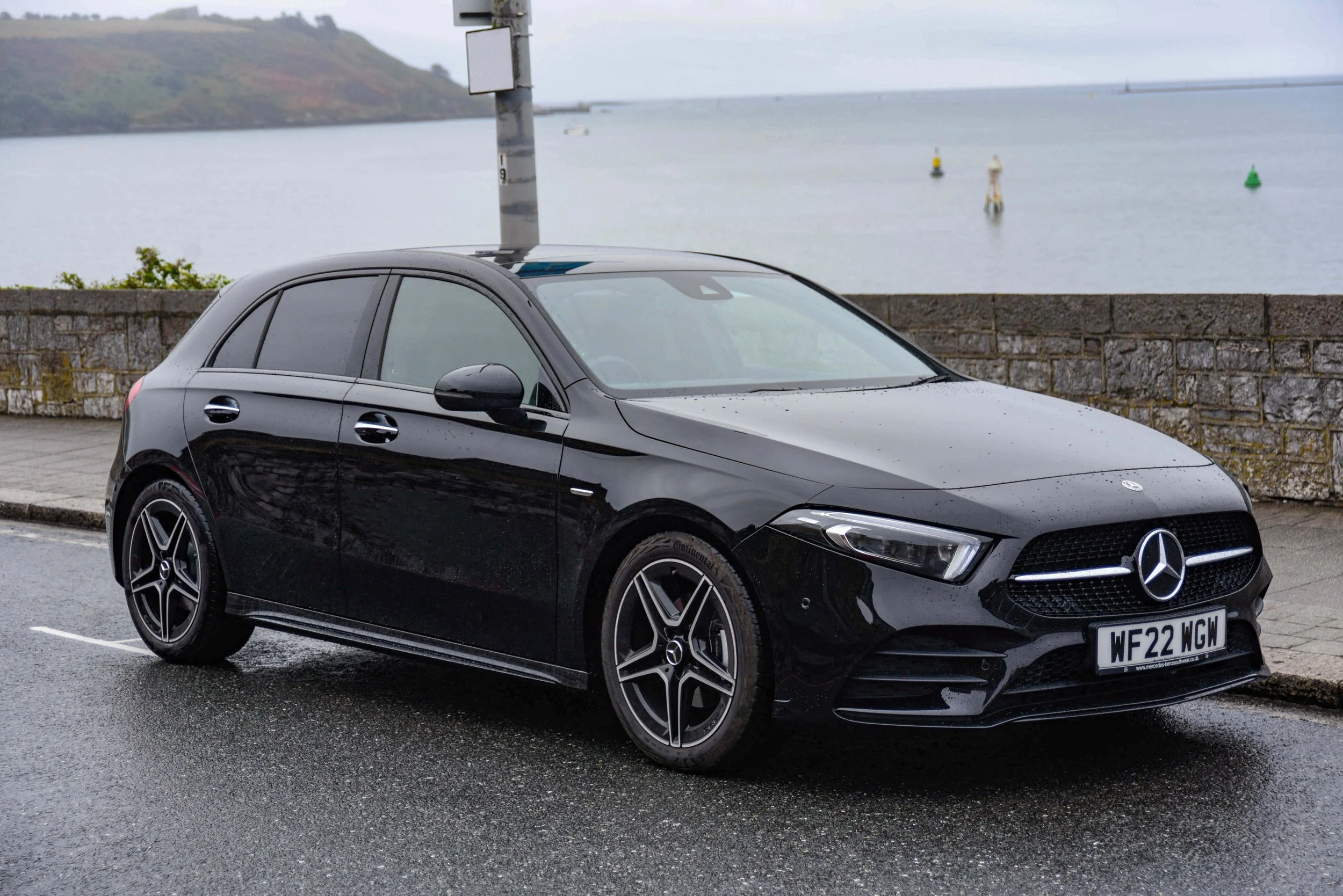 A sleek black Mercedes-Benz hatchback parked on a wet pavement beside a stone wall. The background features a calm body of water and a cloudy sky.