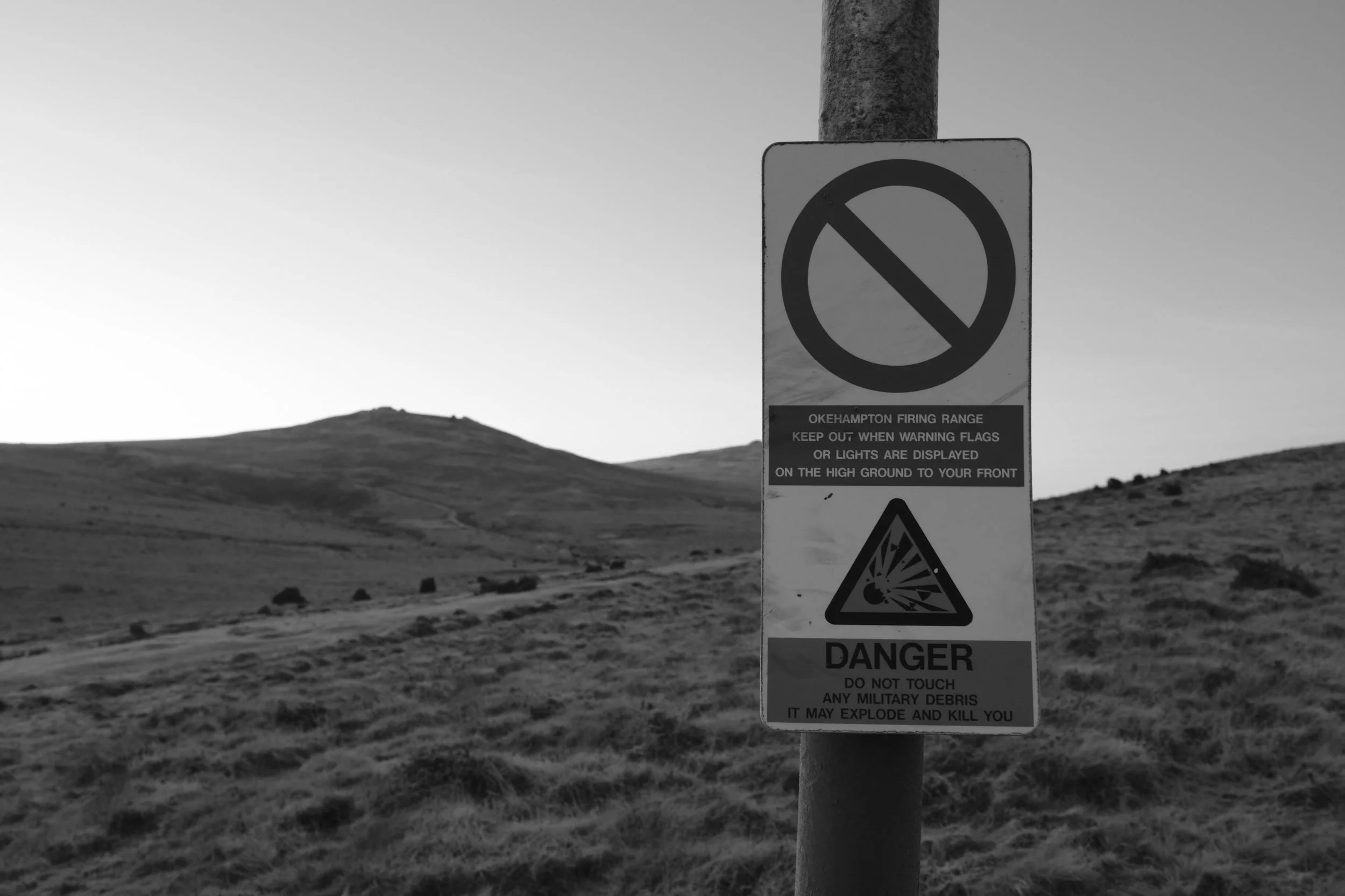 Black and white image of a warning sign on a post in a grassy landscape, indicating a firing range with a danger symbol, emphasizing caution and risk.