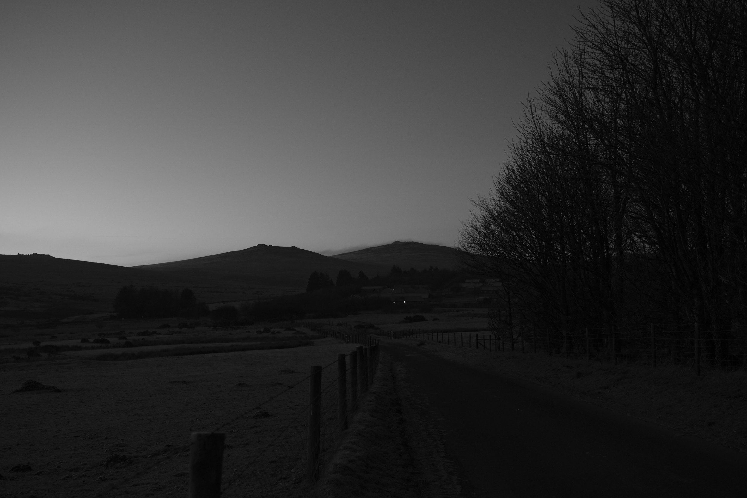 A silhouetted rural landscape at dusk, featuring a winding road lined with bare trees, hills in the background, and a serene, tranquil atmosphere.