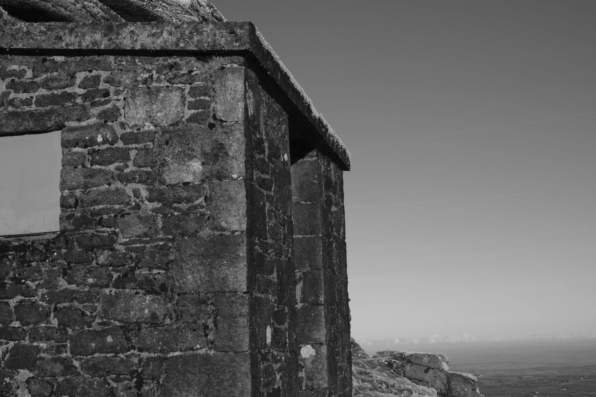 Black and white image of a weathered stone structure with a flat roof, partially covering a window. It's set against a vast, open sky and rocky landscape.