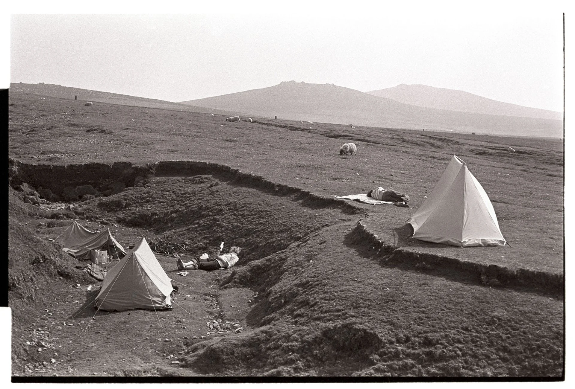 Black and white photograph printed on gelatin paper from James Ravilious of Dartmoor National Park. People sunbathe in the foregrount with two tents errected and sheep in the background as well as Yes Tor and Higher Willhays.