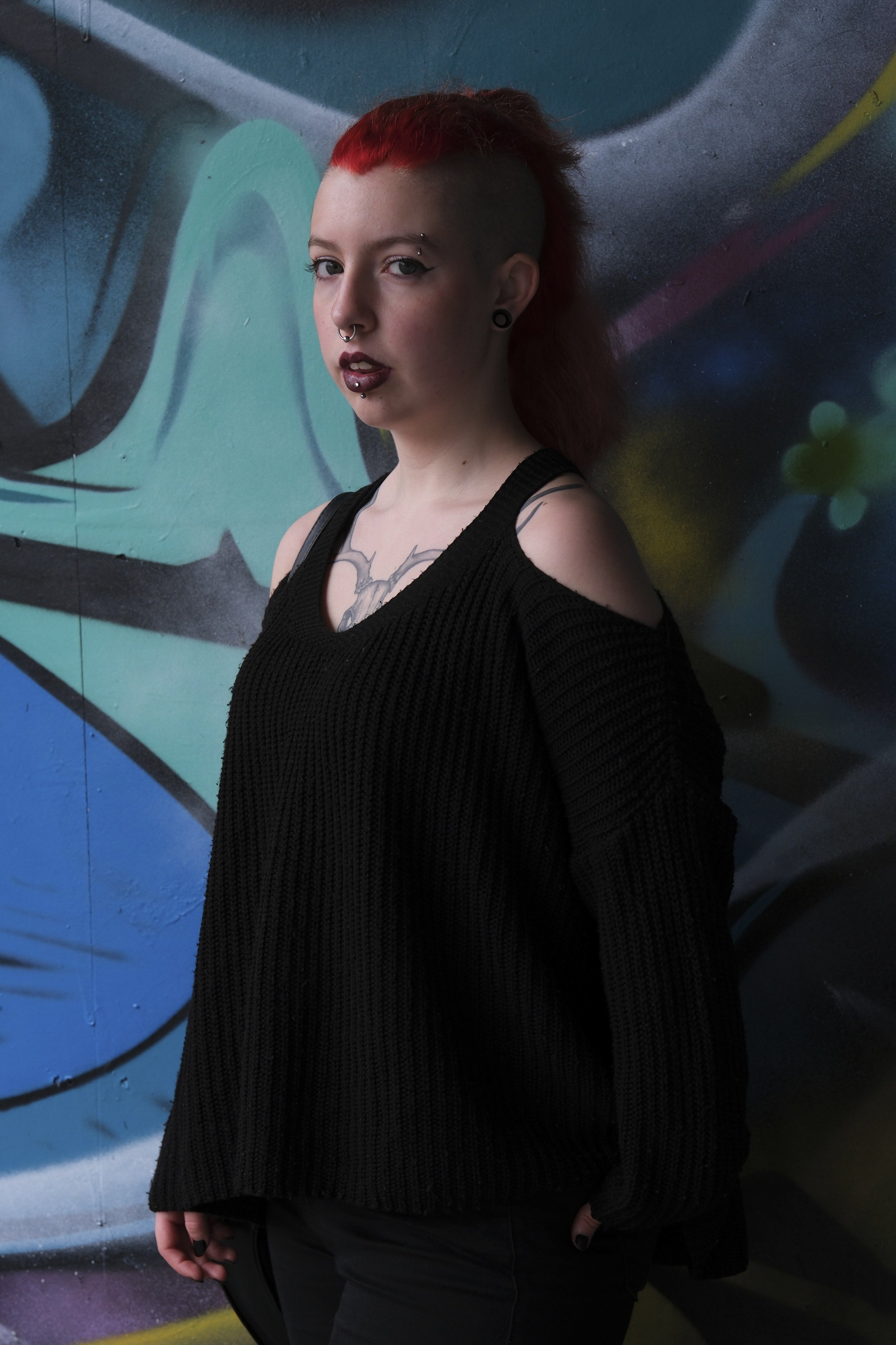 Person with red hair styled in a mohawk stands against a vibrant graffiti backdrop. They wear a black cutout sweater, exuding confidence.