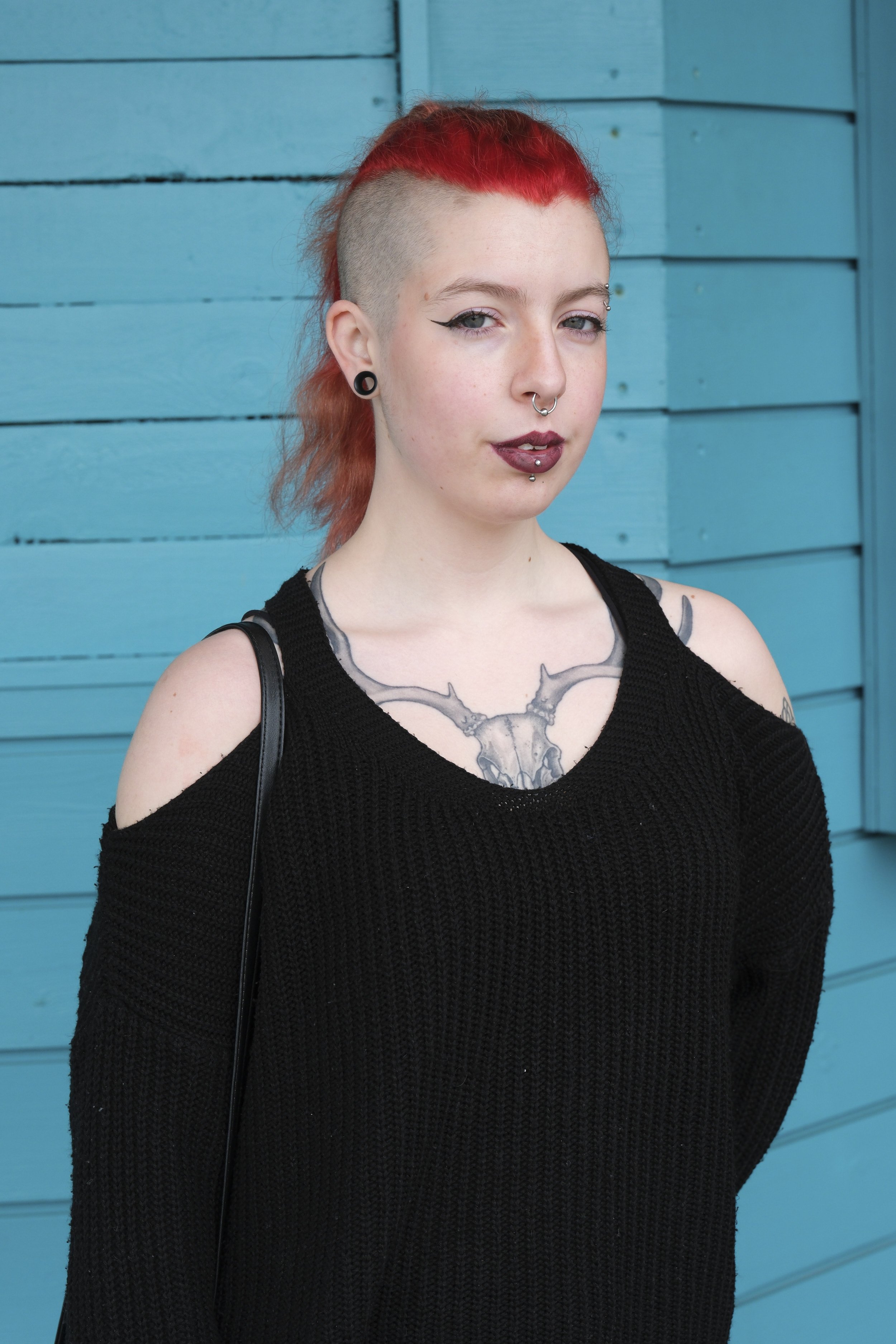 A person with a shaved side and red hair stands confidently against a turquoise wooden wall. She wears a black sweater, showing tattoos and piercings.