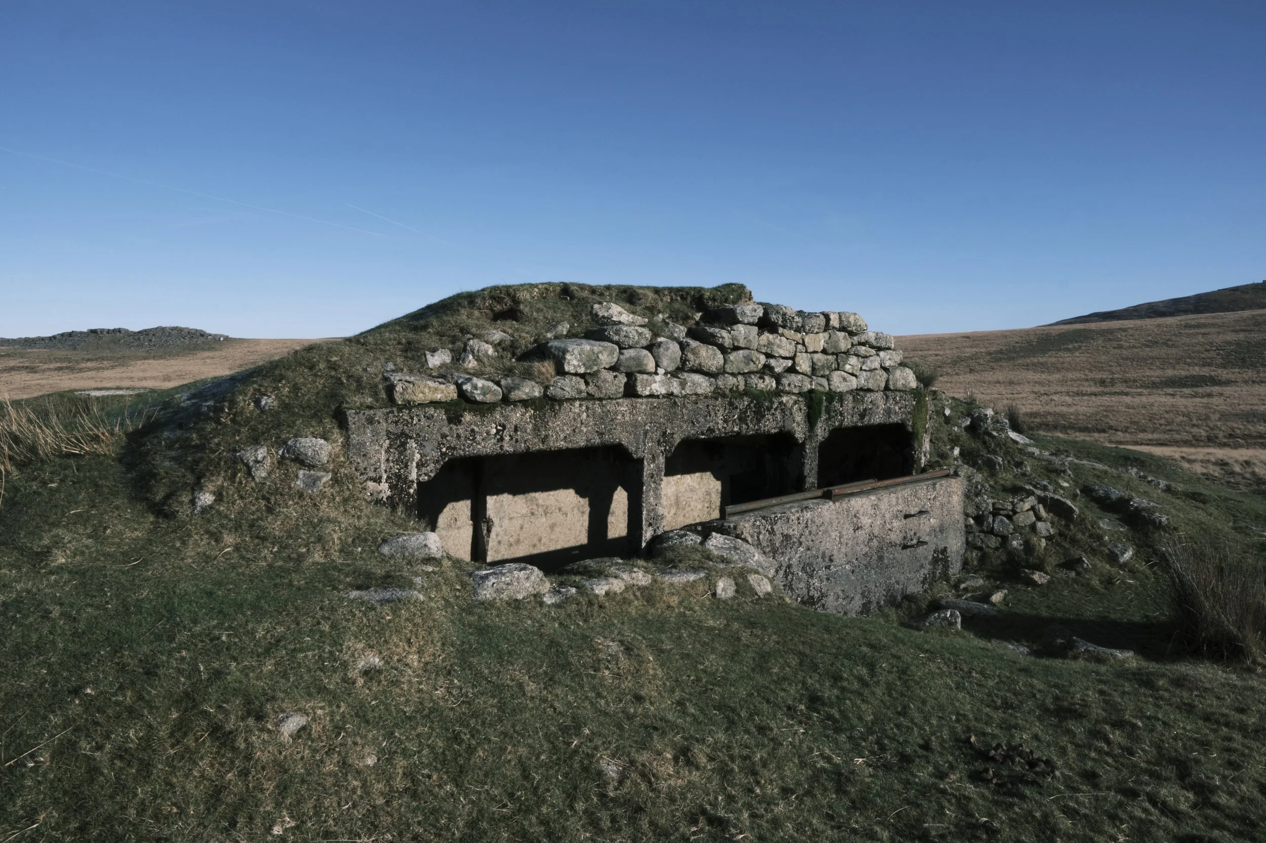 Bunker with stone walls and grassy top in a vast, open landscape under a clear blue sky. The scene conveys isolation and historical intrigue.