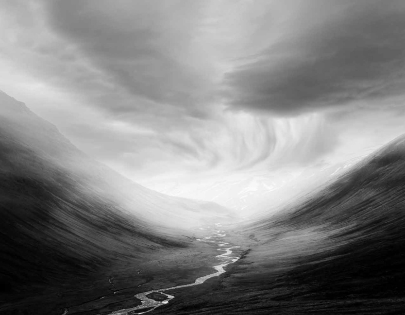Black and white photograph depicting a stream leading down a valley with blurred mountainous landscape running through either side of the stream