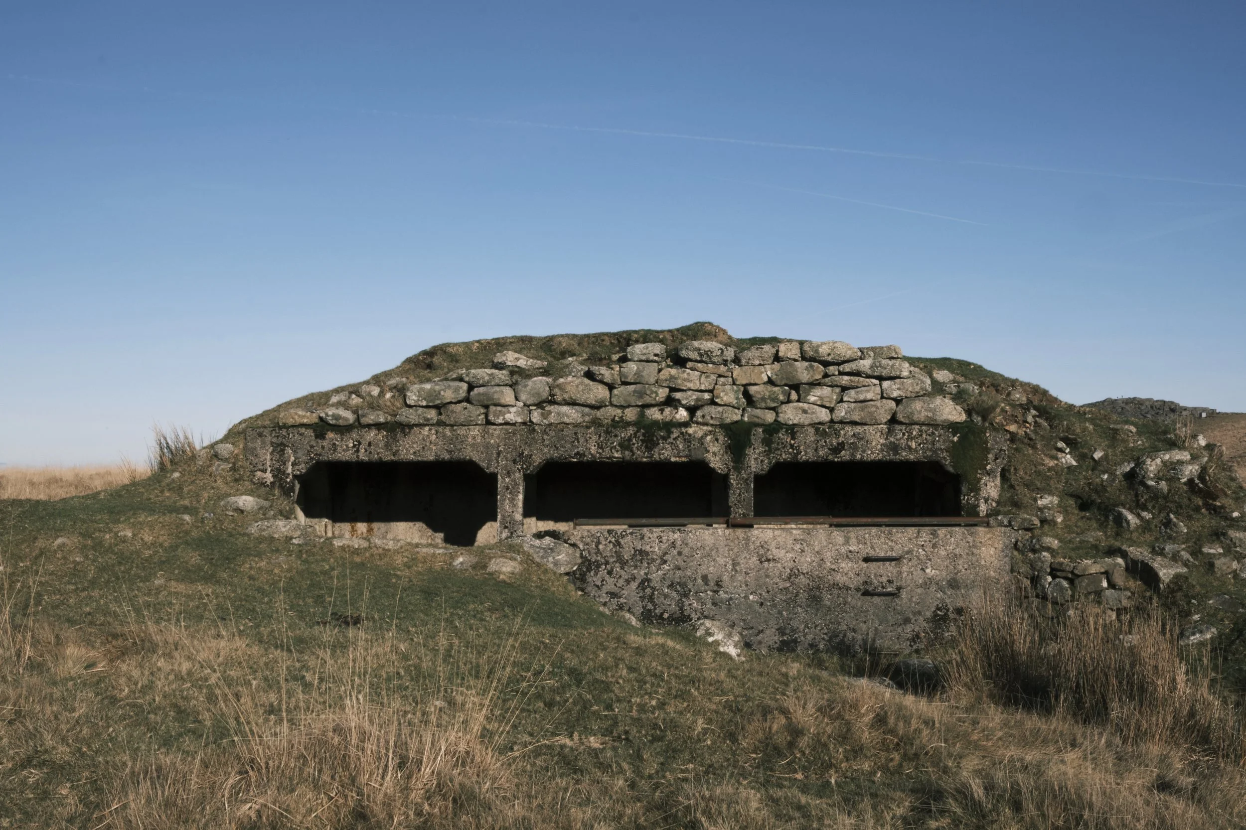 Stone bunker partially buried in grassy terrain under a clear blue sky. The structure conveys a sense of history and solitude in an open landscape.