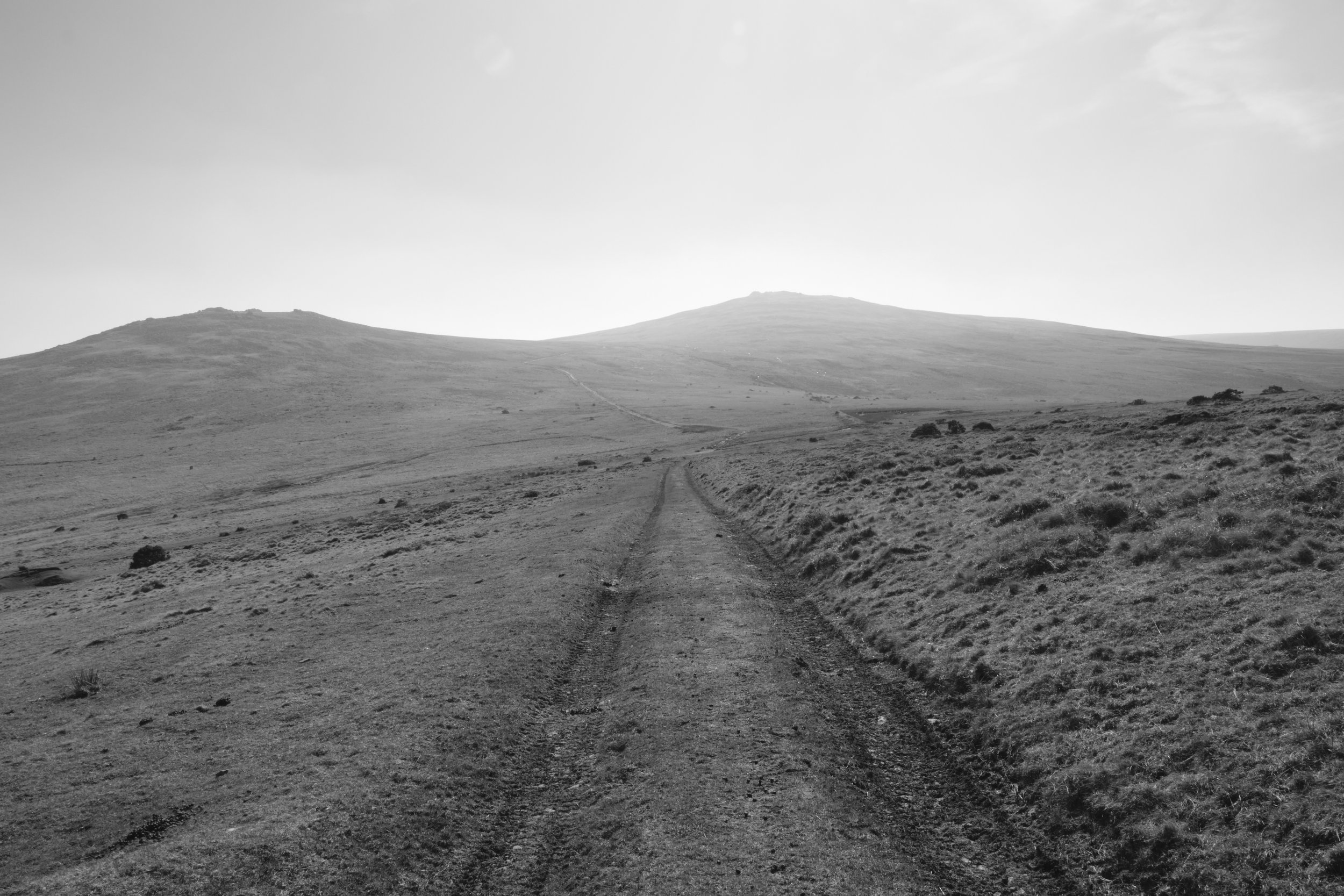 Black and white photograph in Okehampton, Dartmoor National Park. Car tyre tracks are visible against a backdrop of Higher Willhays and Yes Tor in hazy bright conditions. Grass is shown on either side of the tire tracks