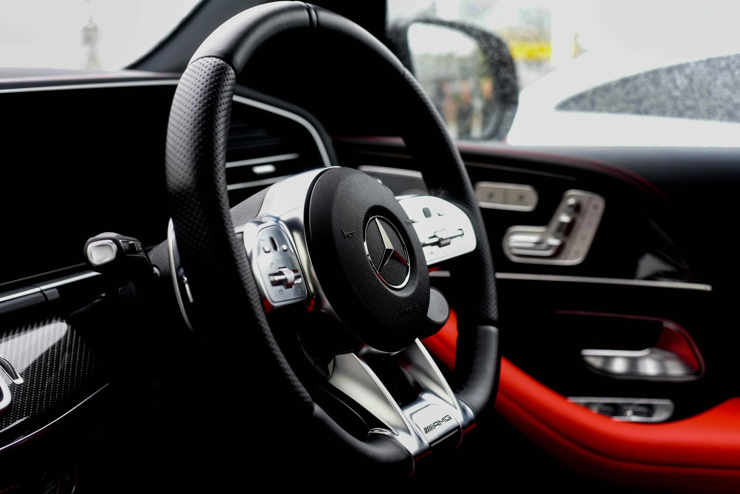 Close-up of a luxury car's interior focusing on a black and silver steering wheel with a logo. Red and black accents create a sleek, modern look.