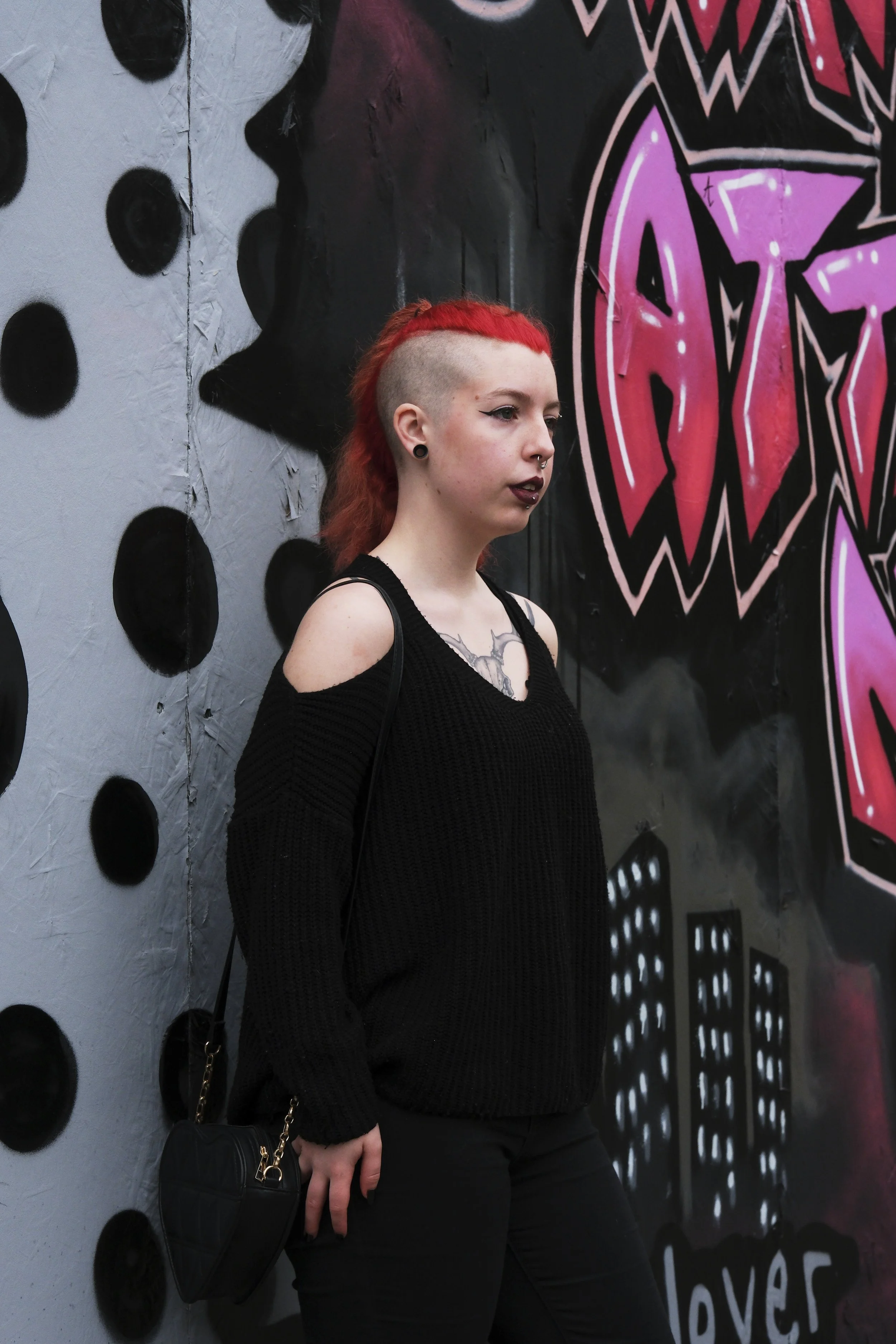 A person with red hair and a side shave stands confidently against a graffiti wall. She wears a black sweater, exuding a bold, urban vibe.