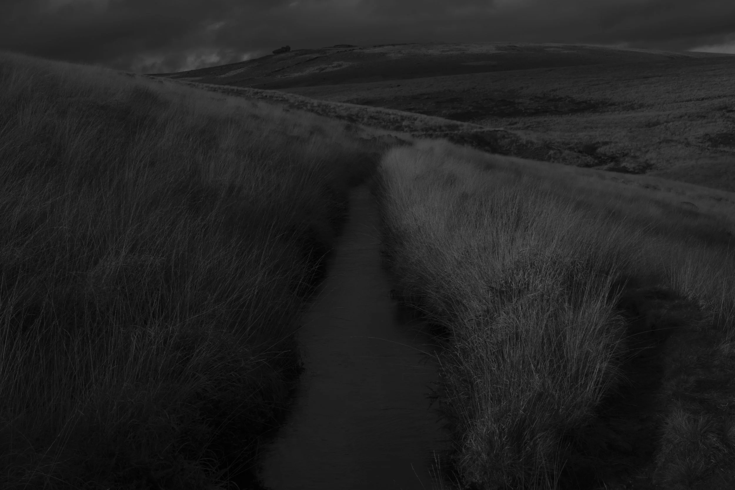 A narrow stream winds through tall grass-covered hills under a cloudy, dark sky, creating a moody and atmospheric landscape.