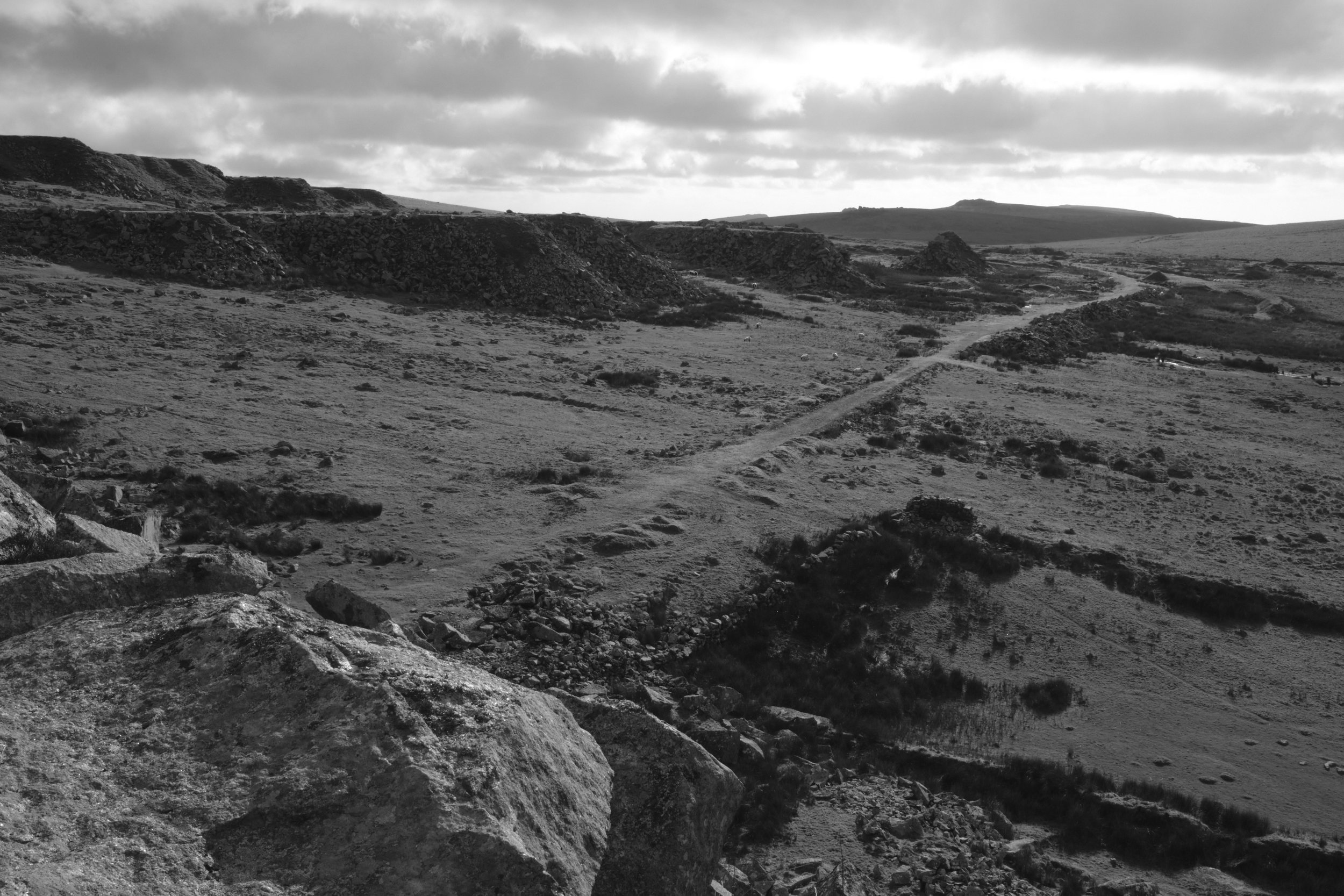 Black and white photo of a rugged, arid landscape with rolling hills, rocks, and a dirt path winding through the terrain, under a cloudy sky.