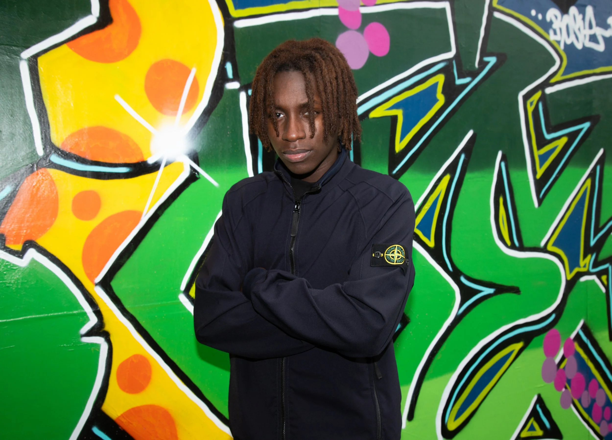 A Colour photograph of a man wearing a black Stone Island jacket with green and yellow graffiti in the background.
