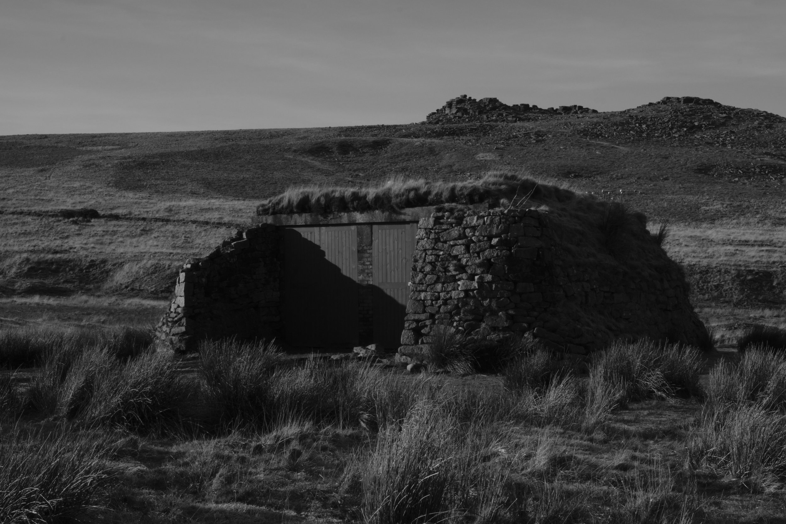 A stone hut with a grassy roof stands isolated in a barren landscape. The scene is monochromatic, conveying a somber, timeless atmosphere.