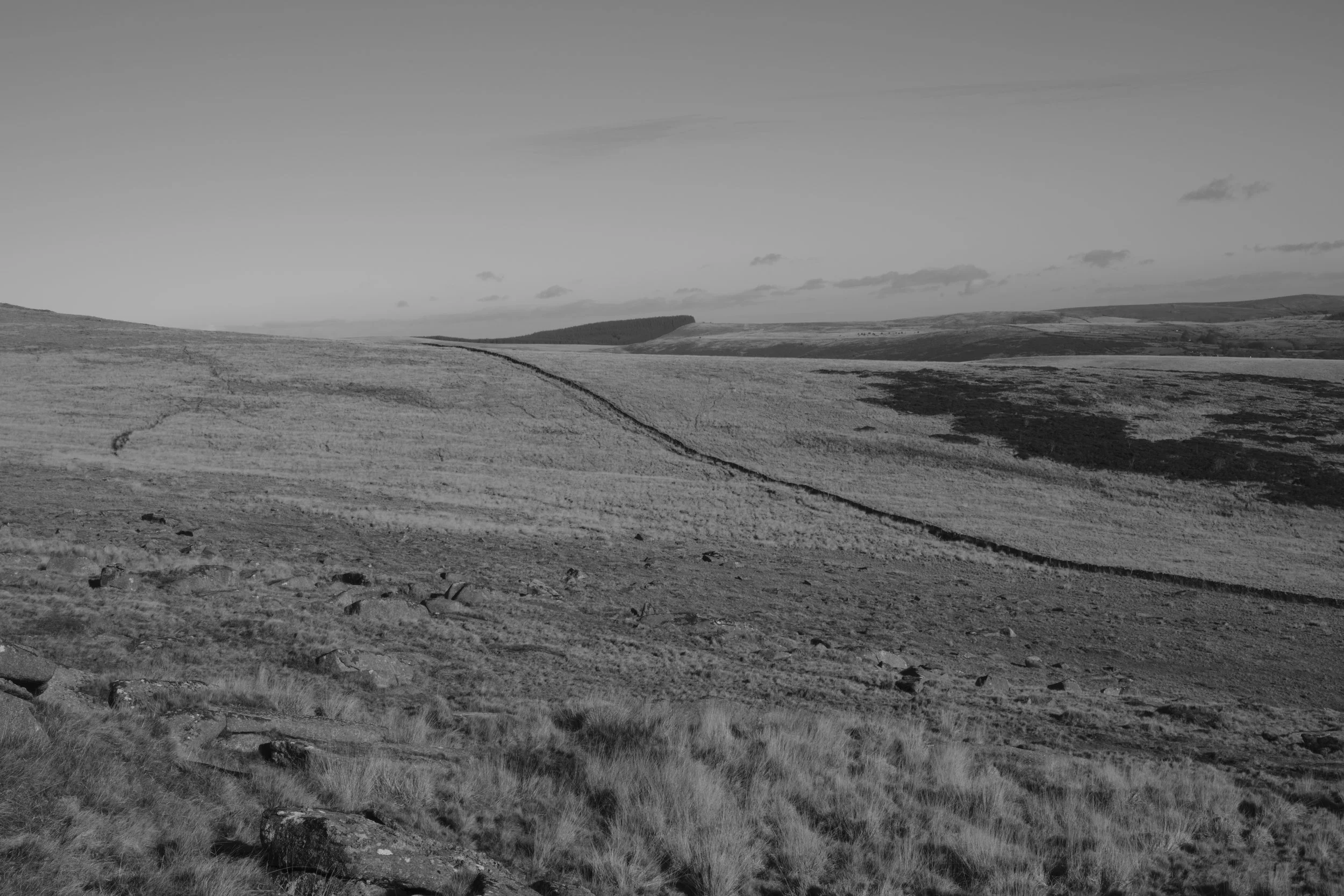 A vast, monochrome landscape features rolling hills and a stone wall stretching toward the horizon under a clear sky, evoking solitude and tranquility.