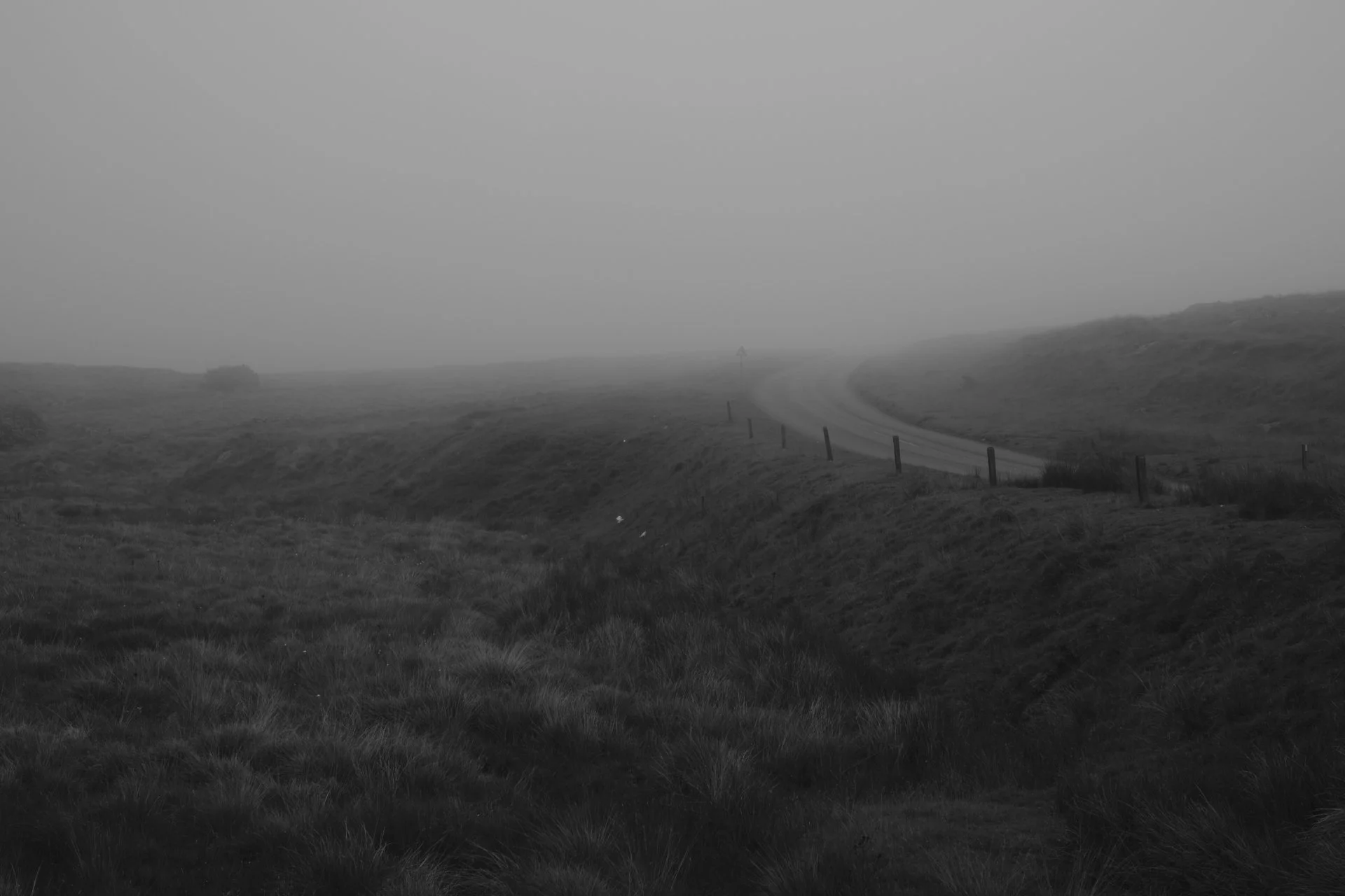 Misty landscape in grayscale, featuring a winding road disappearing into foggy distance. Grass and hills create a serene, somber atmosphere.