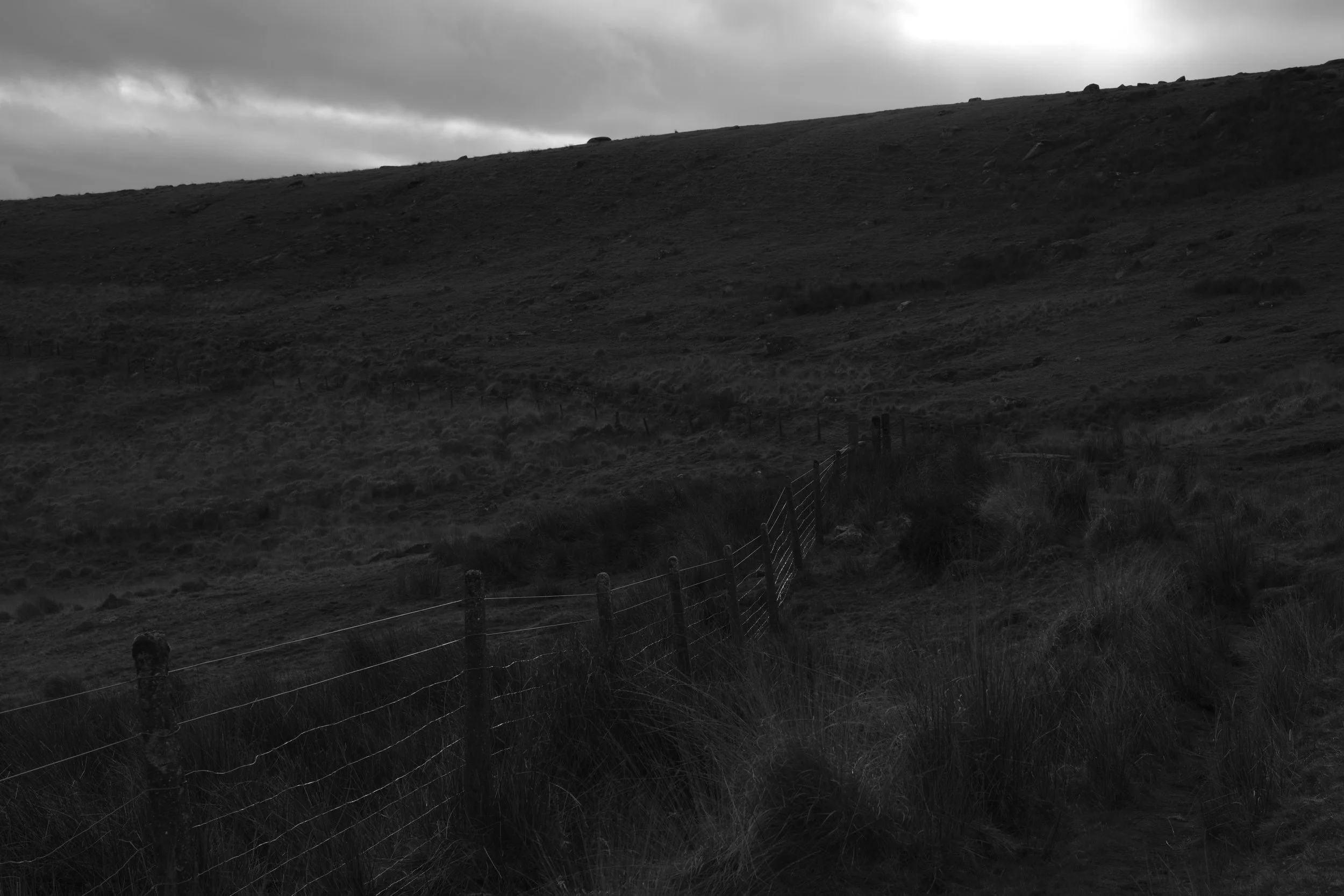 A dark hillside landscape under an overcast sky, with a wire fence extending into the distance. The scene conveys a moody and desolate atmosphere.