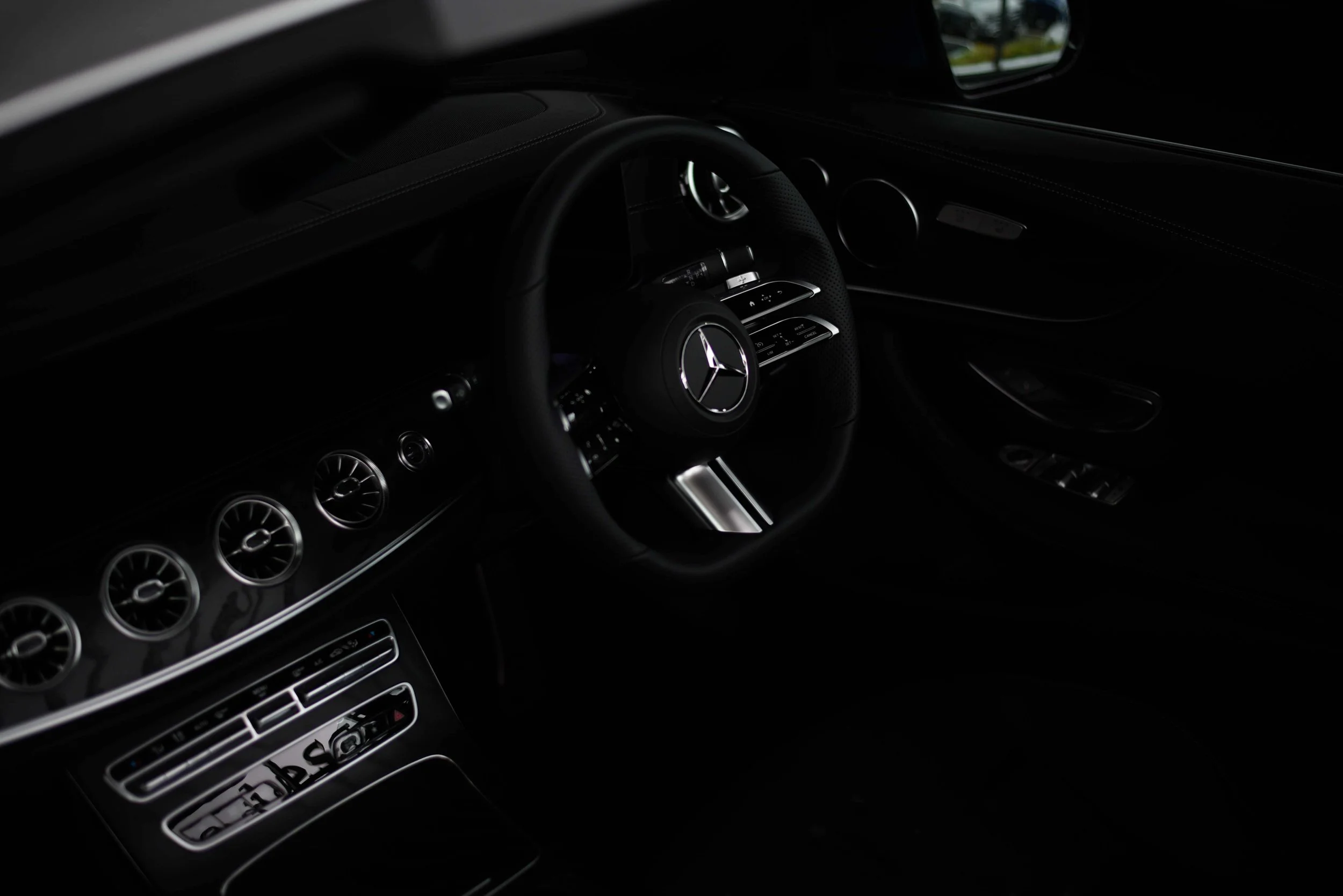 Interior of a luxury car with a black steering wheel featuring a Mercedes logo, sleek dashboard with chrome accents, and air vents. The atmosphere is dark and sophisticated.