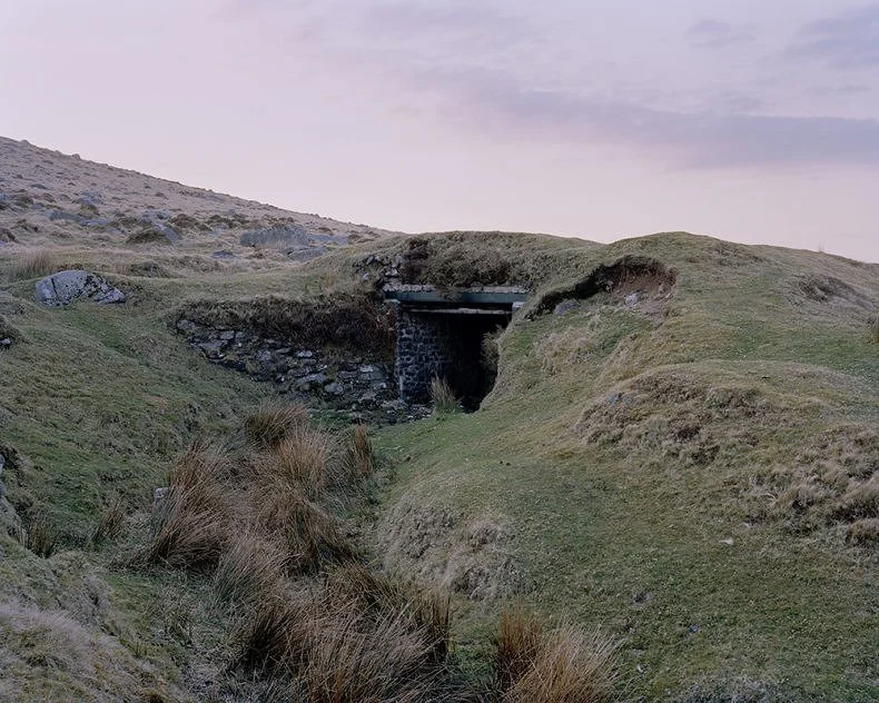 Colour photograph of Dartmoor National Park. A military storage unit is seen in rocky terrain with green grass, some of it overgrown, and rocks, especially on the left side of the image.