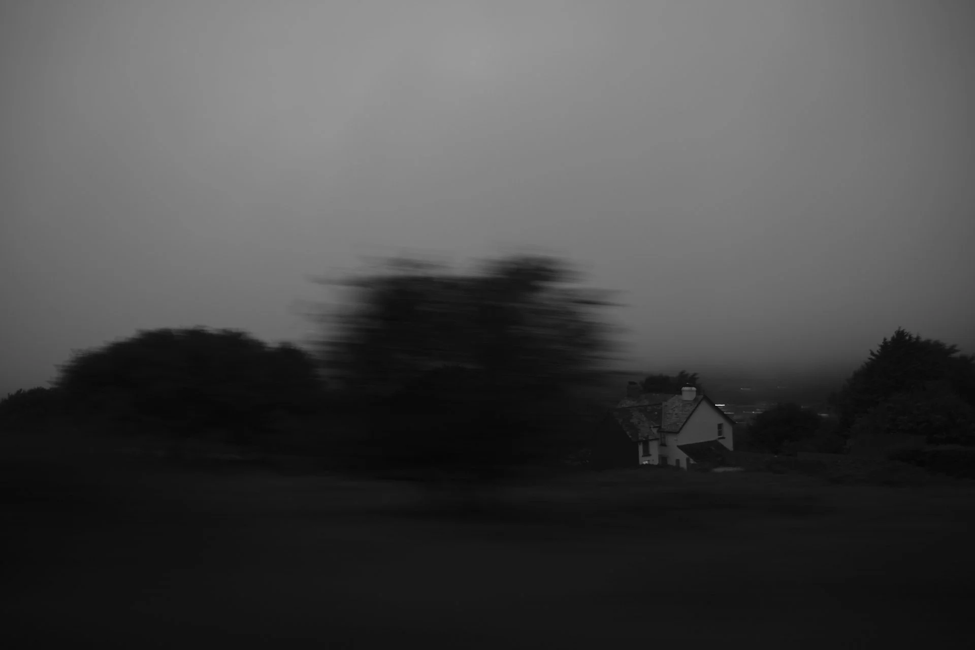 A black and white photo of a rural landscape with a house, trees, and open land, taken in low light or during dusk.