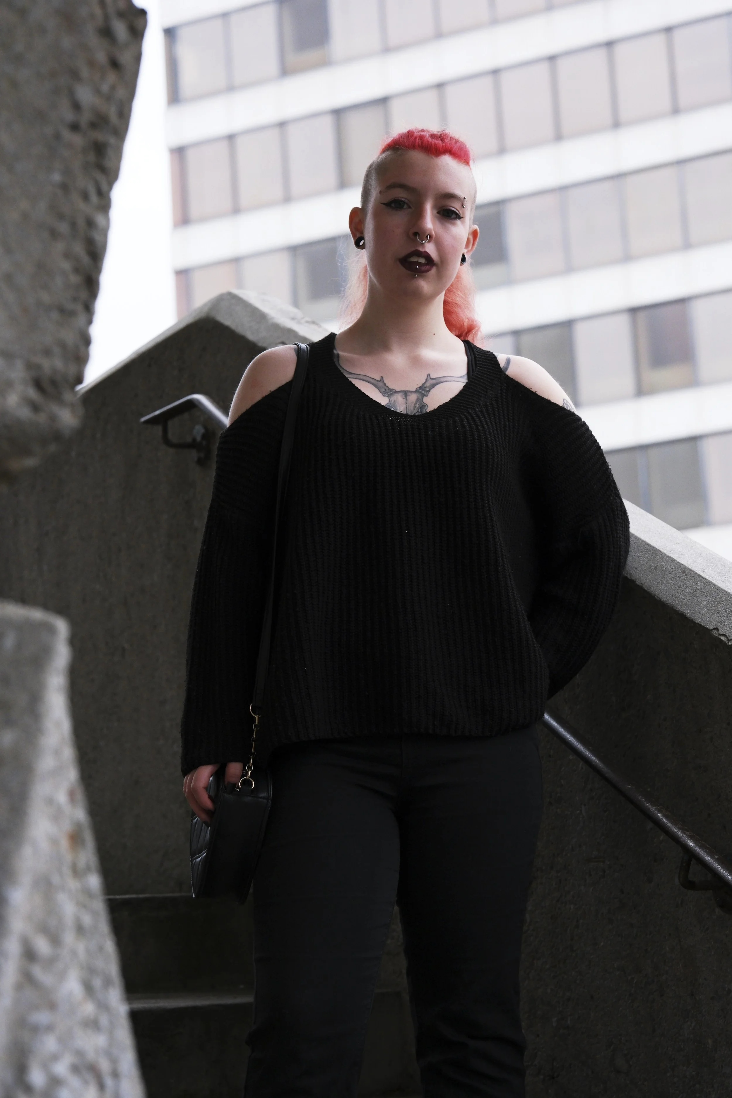 A person with vibrant pink hair, wearing a black outfit, stands confidently on a concrete staircase. An office building forms the background.